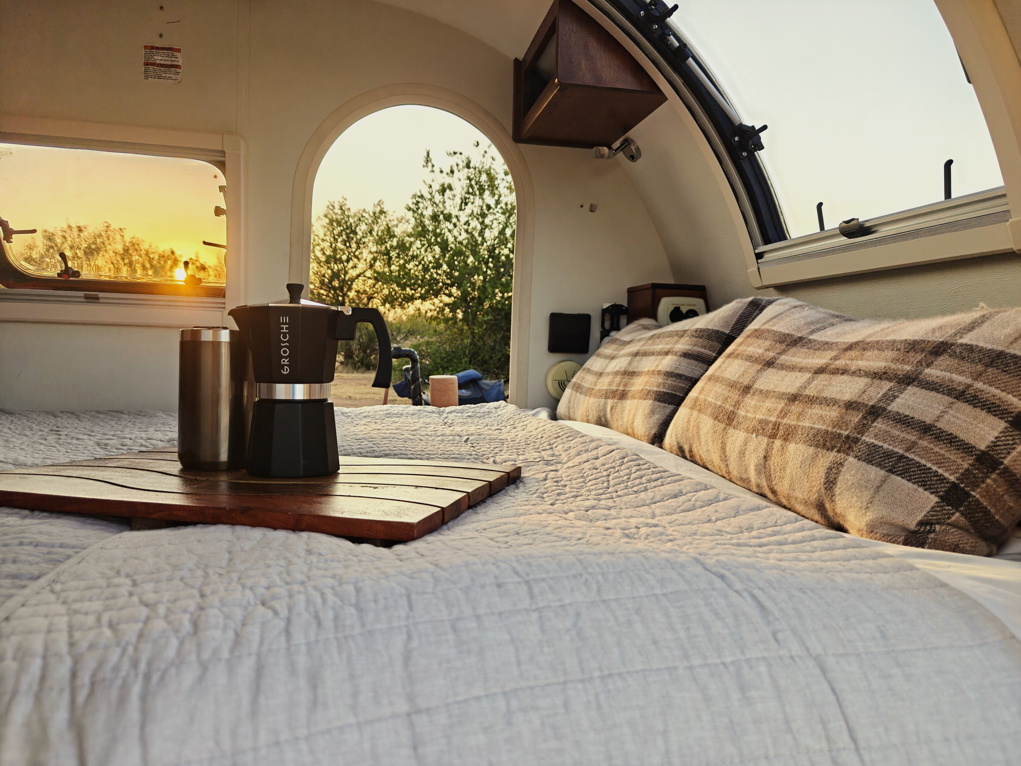 Cozy RV rental interior with a Moka pot & coffee mug on the bed, sun setting through the window. Relaxing travel.. nuCamp T@G 2018