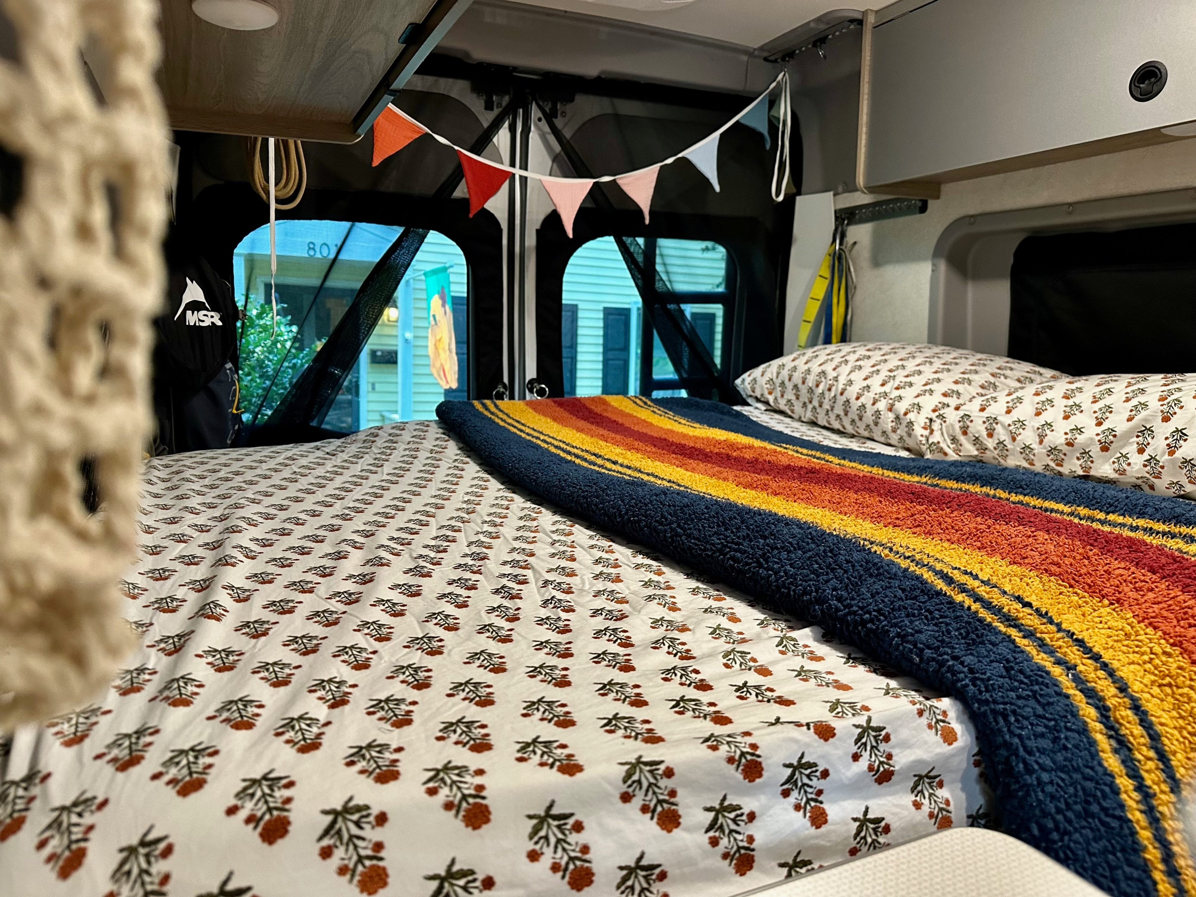 Cozy RV rental bed with floral sheets, a striped blanket, and colorful pennants. A glimpse of house "801" is visible outside.. Winnebago Solis 2023