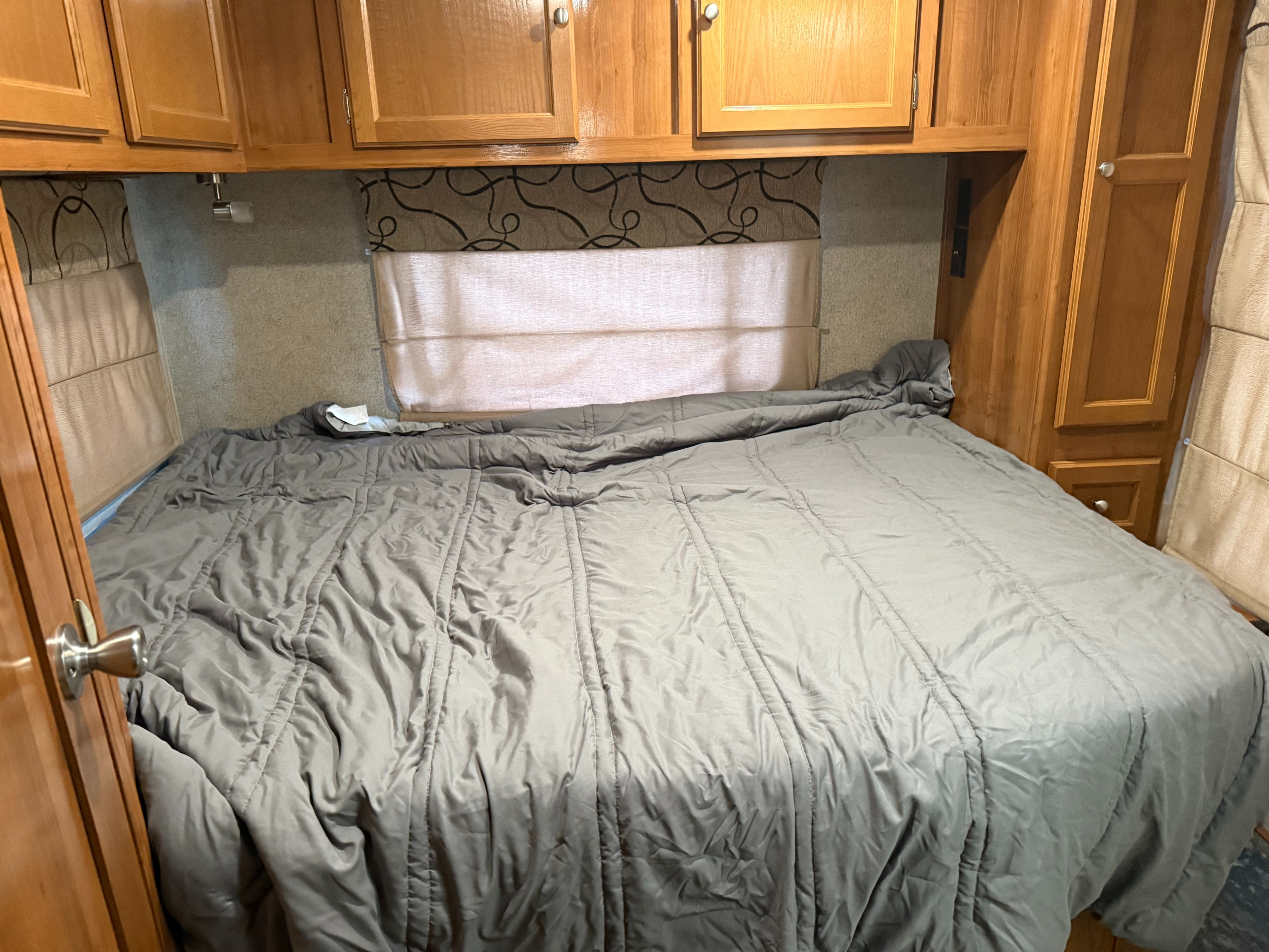 Cozy RV rental bedroom featuring a large bed with a grey comforter, wooden overhead cabinets, and window shades.. Coachmen Leprechaun 2019