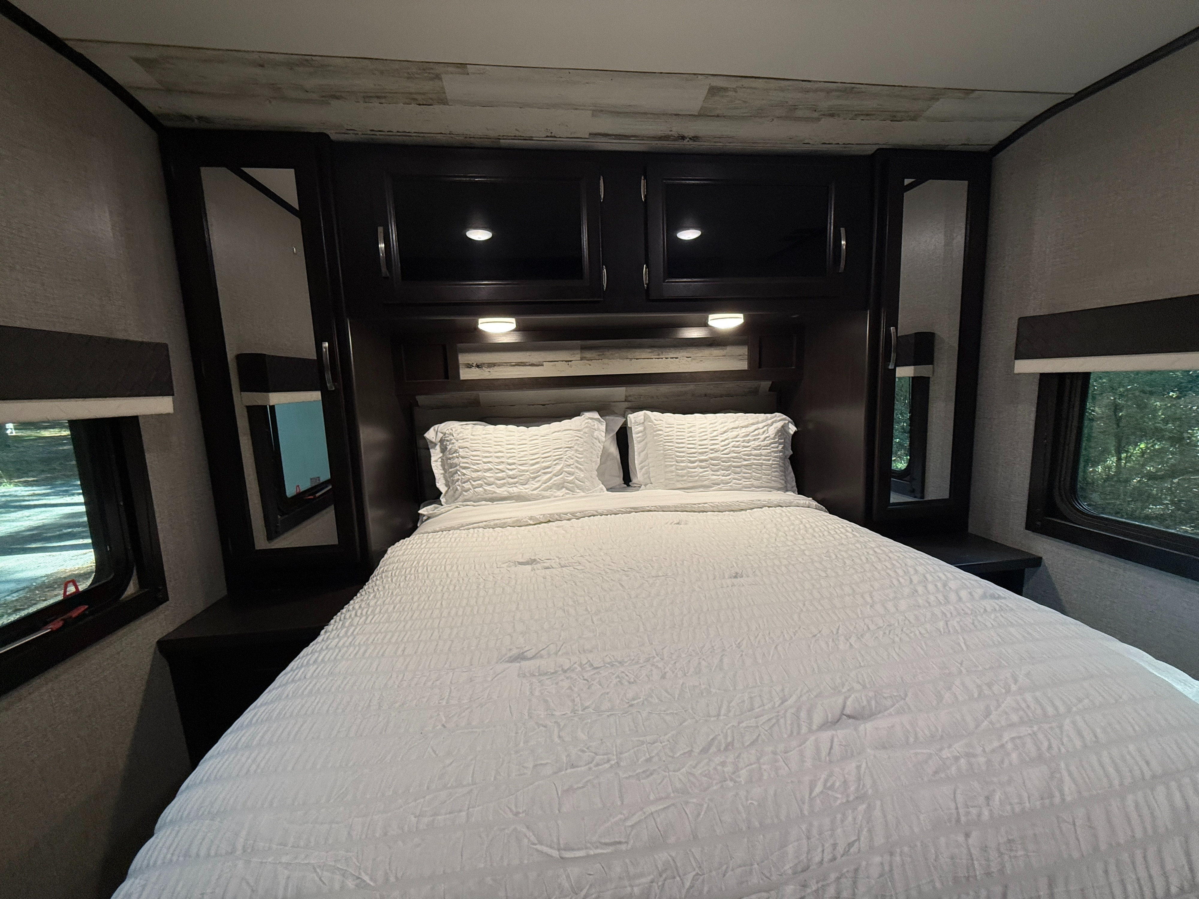 Cozy RV rental bedroom features a comfortable bed with white bedding, overhead cabinets, and side windows with natural views.. Jayco Jay Feather 2020