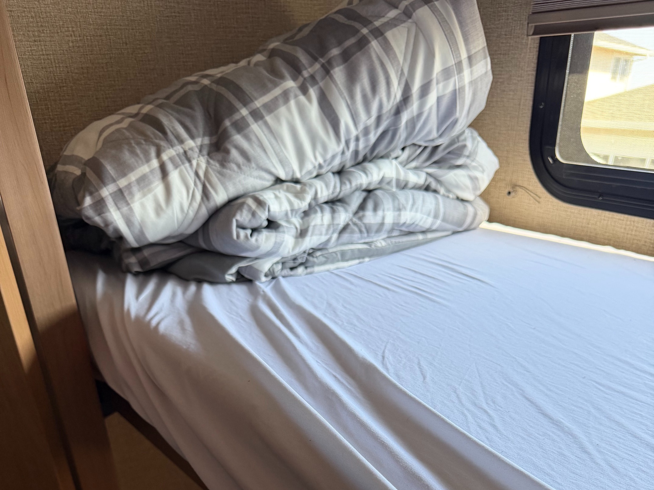 Inside an RV rental: a neatly made bed with a white sheet and rolled gray plaid comforter by the window.. Thor Windsport 2018
