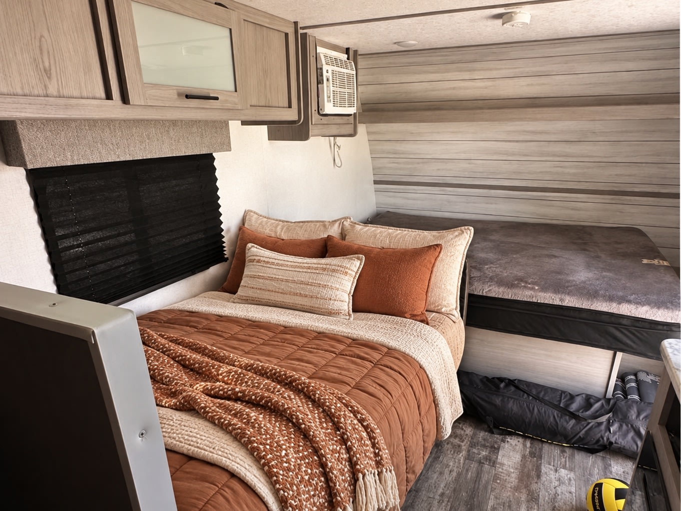 Cozy RV rental bedroom features a bed with rust-orange bedding, knit throw, overhead cabinets, and an upper bunk bed.. Coleman Lantern LT 2024