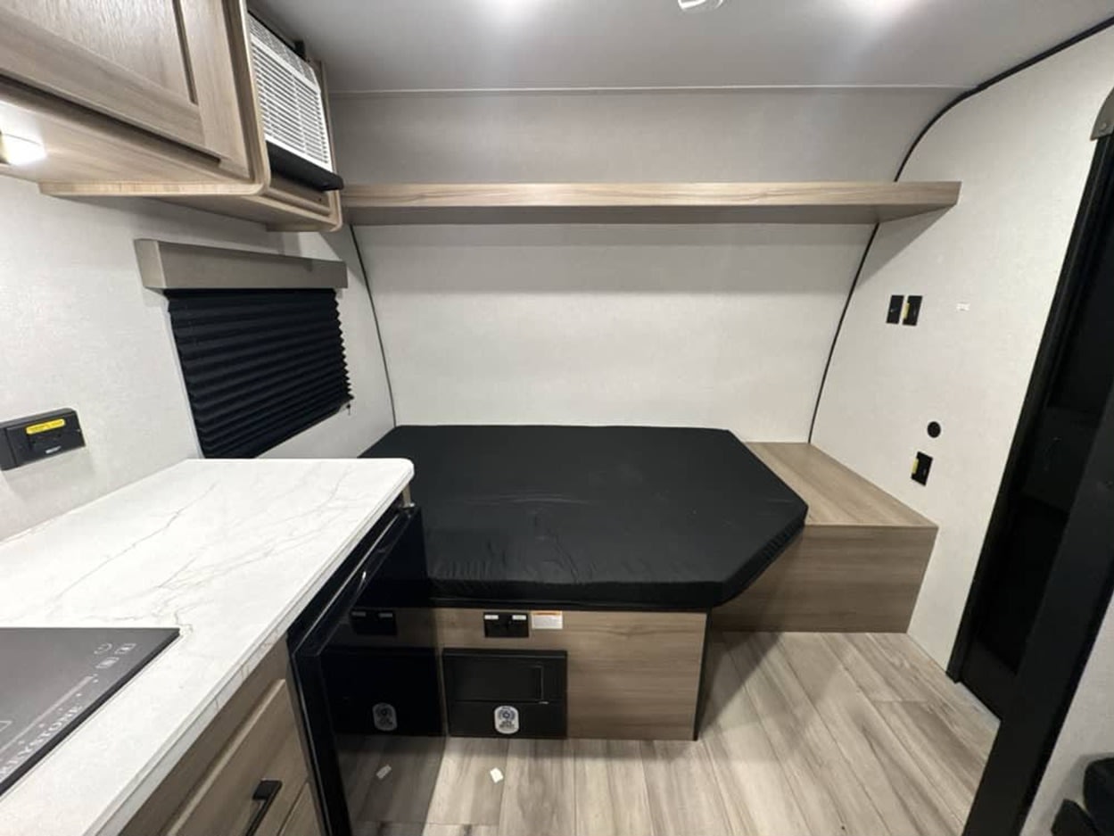 This RV rental interior features a cozy, compact bed, a modern kitchen counter with a stovetop, and overhead storage.. Keystone by Coleman 13B 2027