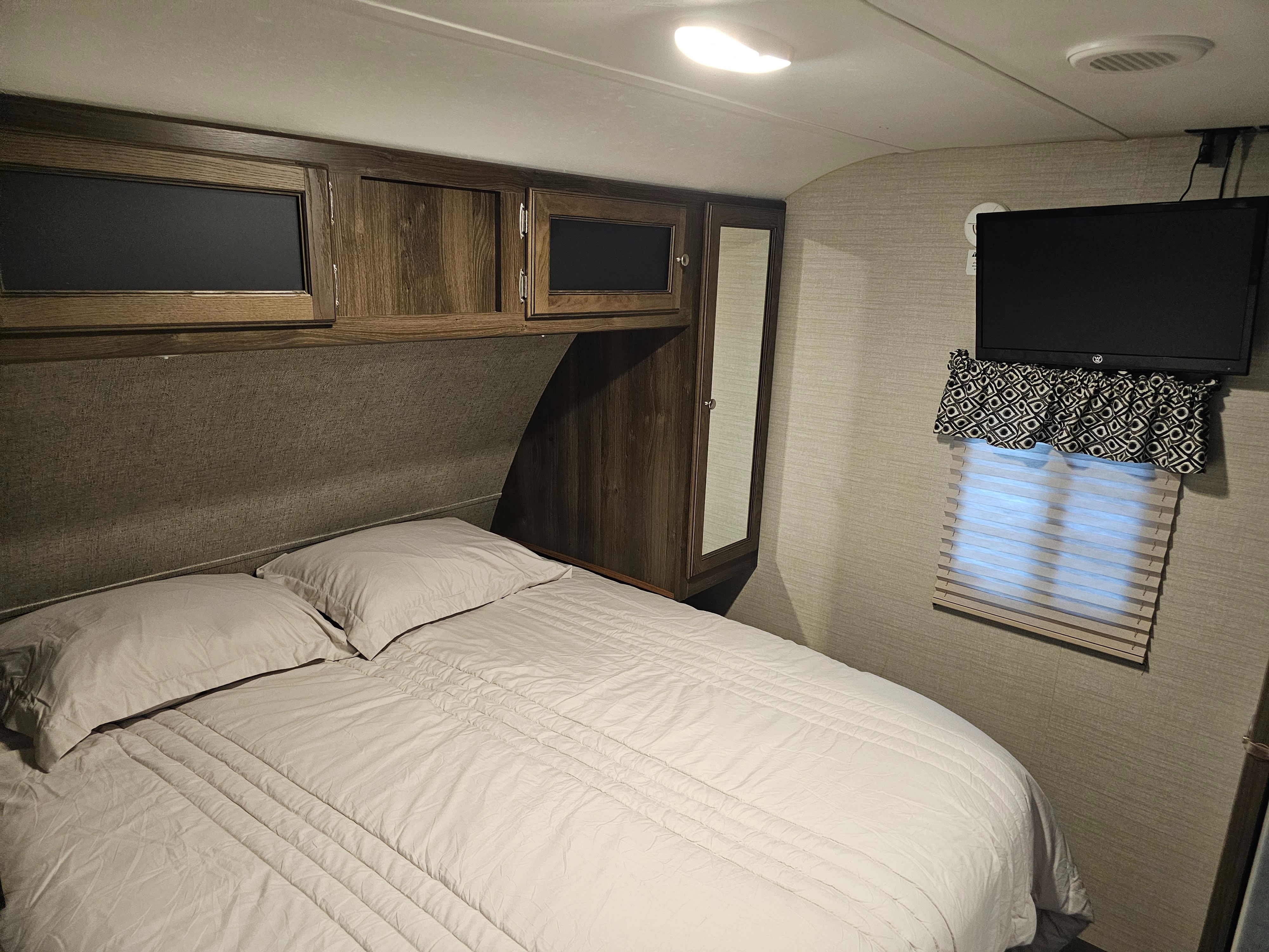 Inside an RV rental, this cozy bedroom features a bed with light bedding, wooden overhead cabinets, a mounted TV, and a window.. Keystone RV Bullet Ultra Lite 2018
