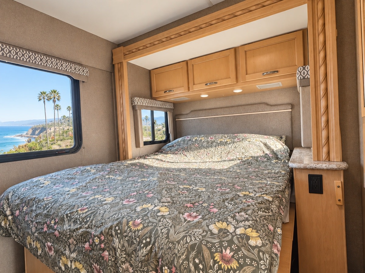 RV rental bedroom featuring a comfy bed with a floral duvet, stunning ocean view, and palm trees from the window.. Thor Freedom Elite 2018