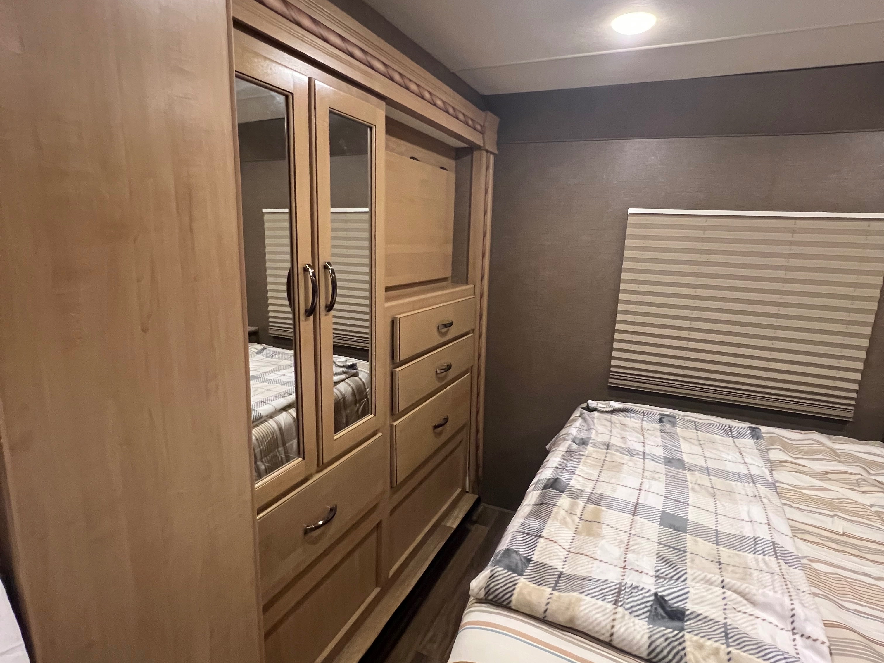 RV rental bedroom featuring a light wood wardrobe with mirrored doors and drawers, a comfy bed, and a pleated window shade.. Thor Four Winds 2018
