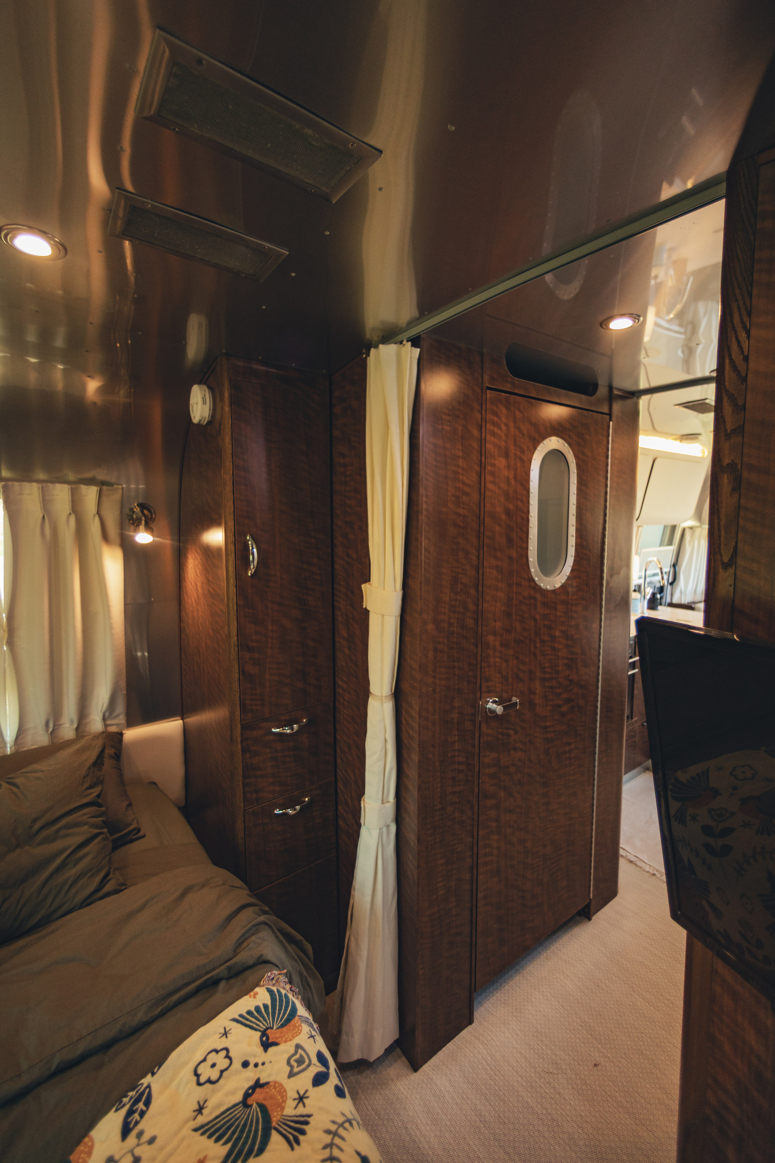 Cozy rv rental bedroom: olive-green bed, bird blanket, dark wood cabinets, metallic ceiling, and a porthole door.. Airstream Globetrotter 2019