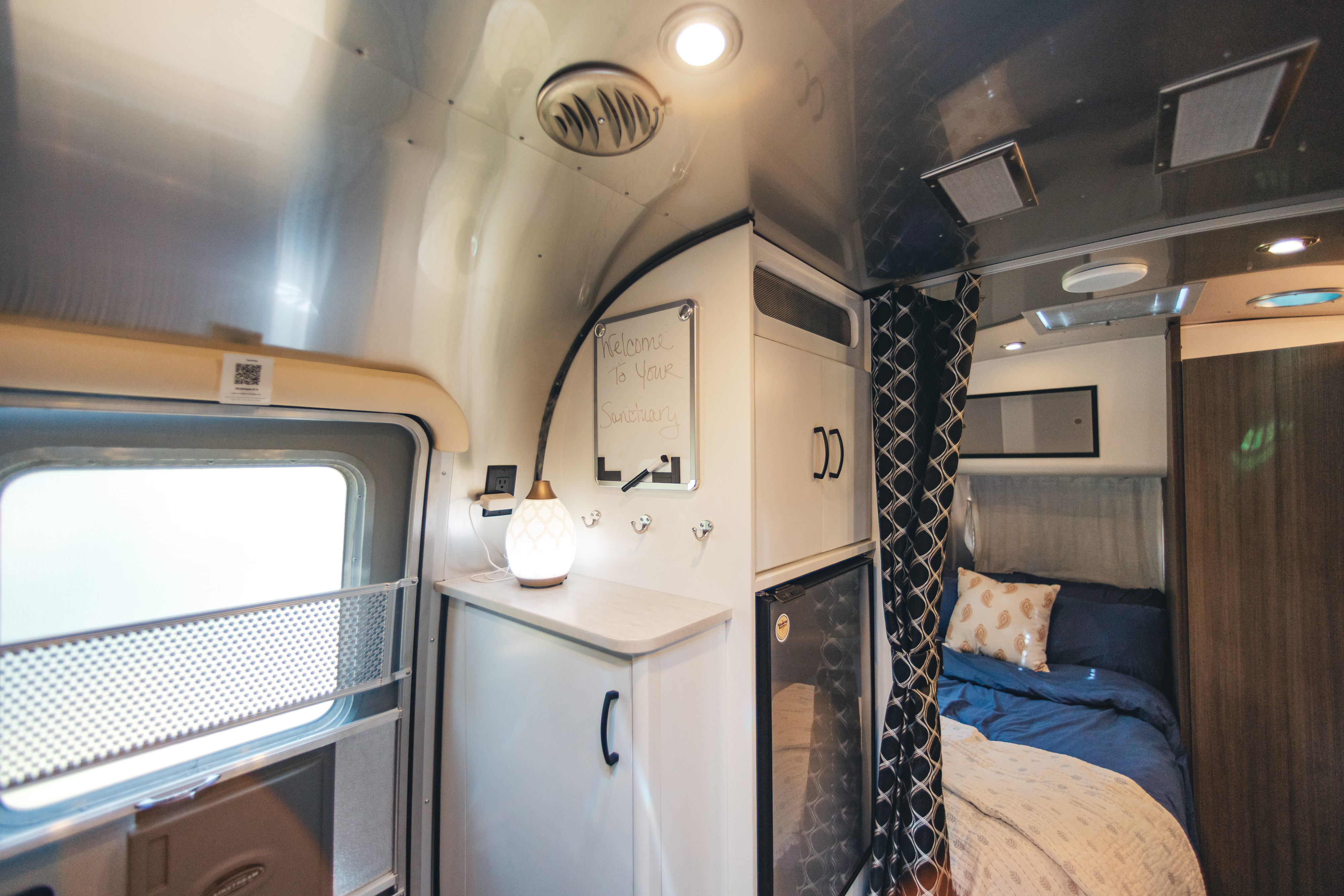 This RV rental interior shows a comfy bed with blue bedding, a counter with a lamp, mini-fridge, and a welcome sign.. Airstream Caravel 2022