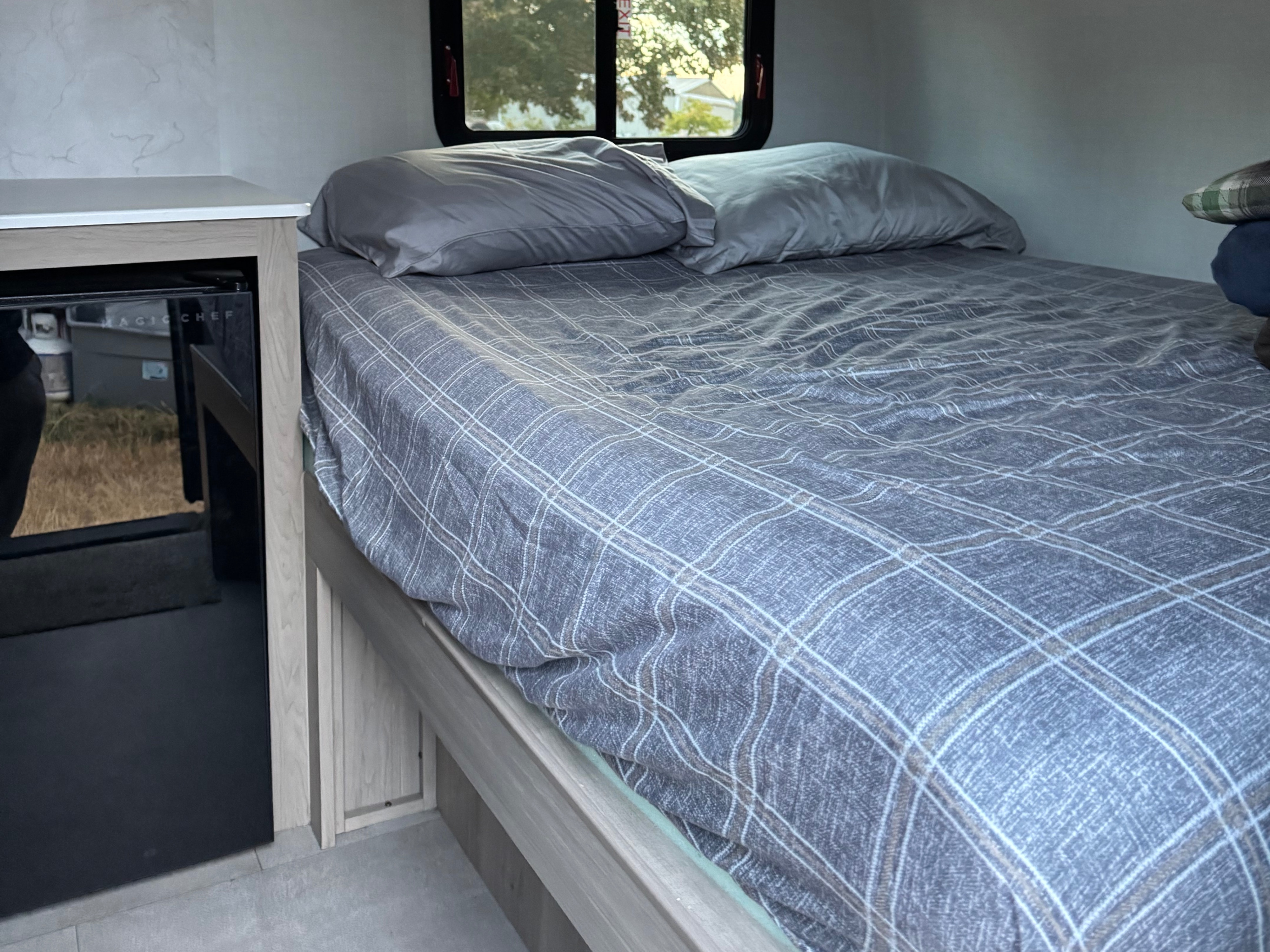 Cozy RV rental interior featuring a bed with gray plaid bedding, two pillows, a window with an outdoor view, and a Magic Chef mini-fridge.. Forest River Rockwood Geopro 2026