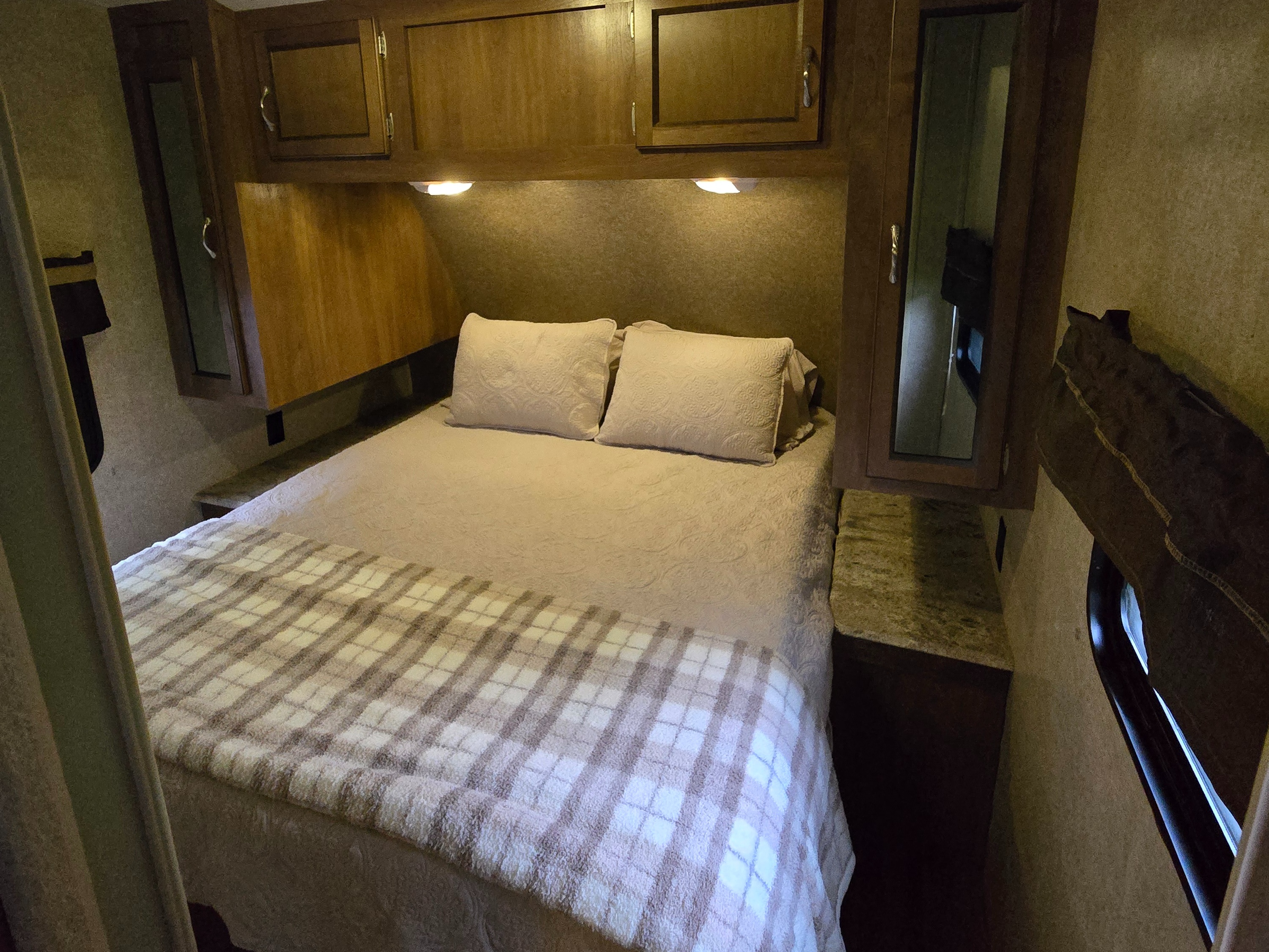 Cozy RV rental bedroom with warm lighting, overhead cabinets, and a comfortable bed with pillows and a plaid blanket.. Coachmen Catalina 2017