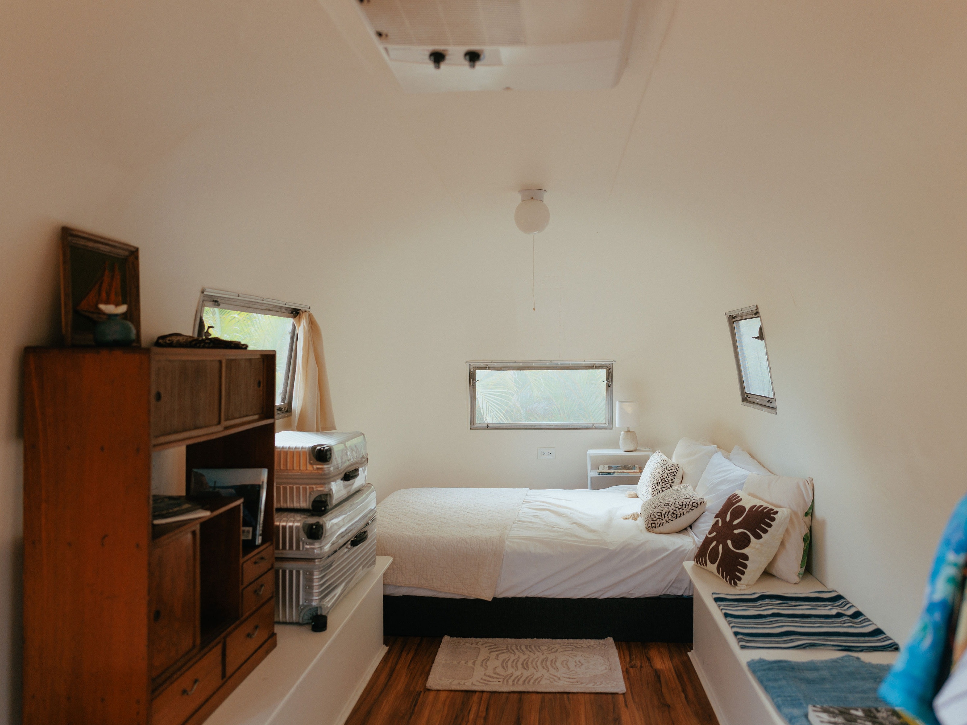This bright RV rental bedroom features a cozy bed with white linens, wooden furnishings, and windows looking out to tropical greenery.. Airstream Land Yacht 1960