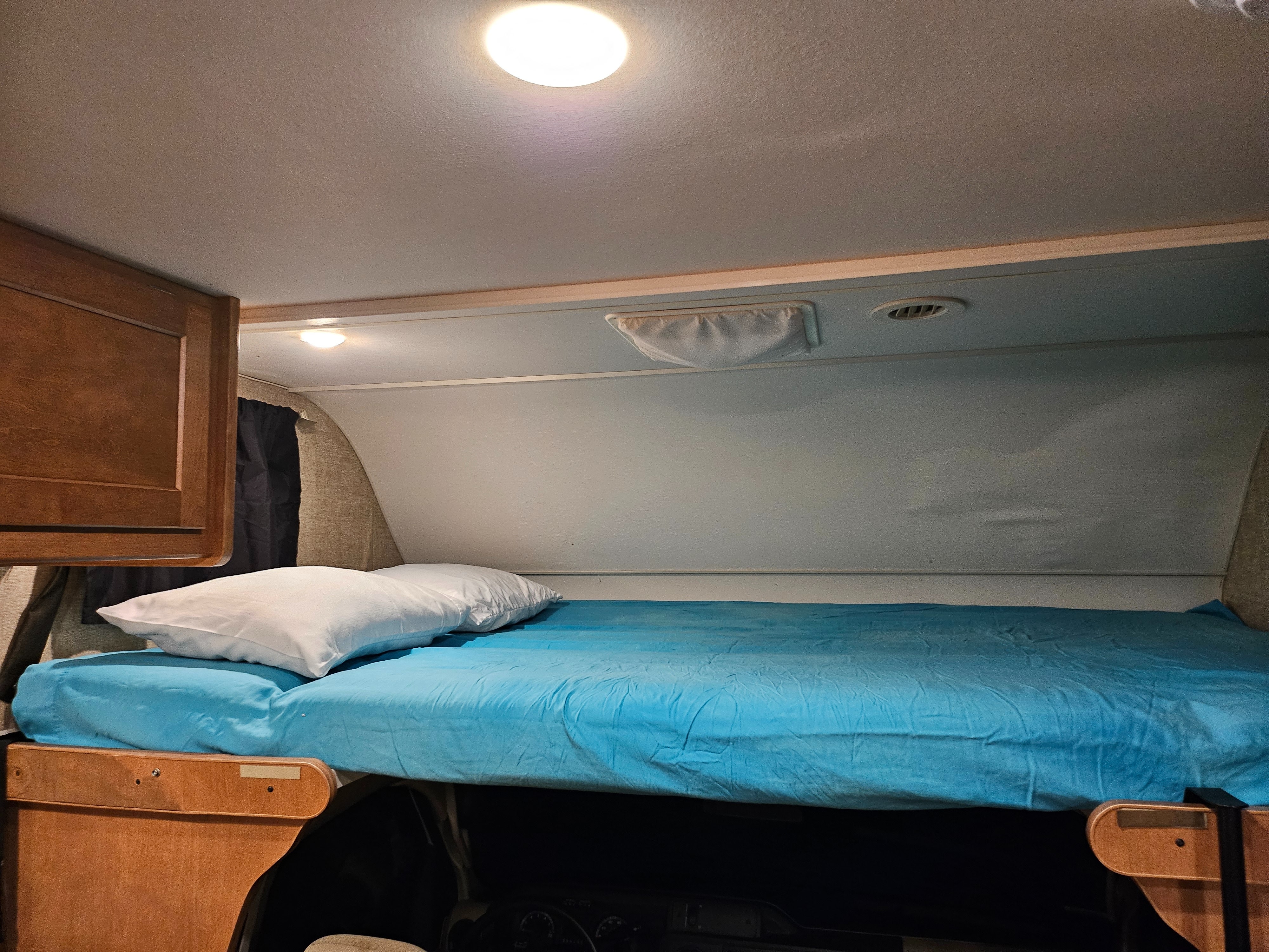 Cozy RV rental sleeping area: a bright bunk with a blue sheet, white pillow, ceiling light, and vent.. Winnebago Minnie Winnie 2018
