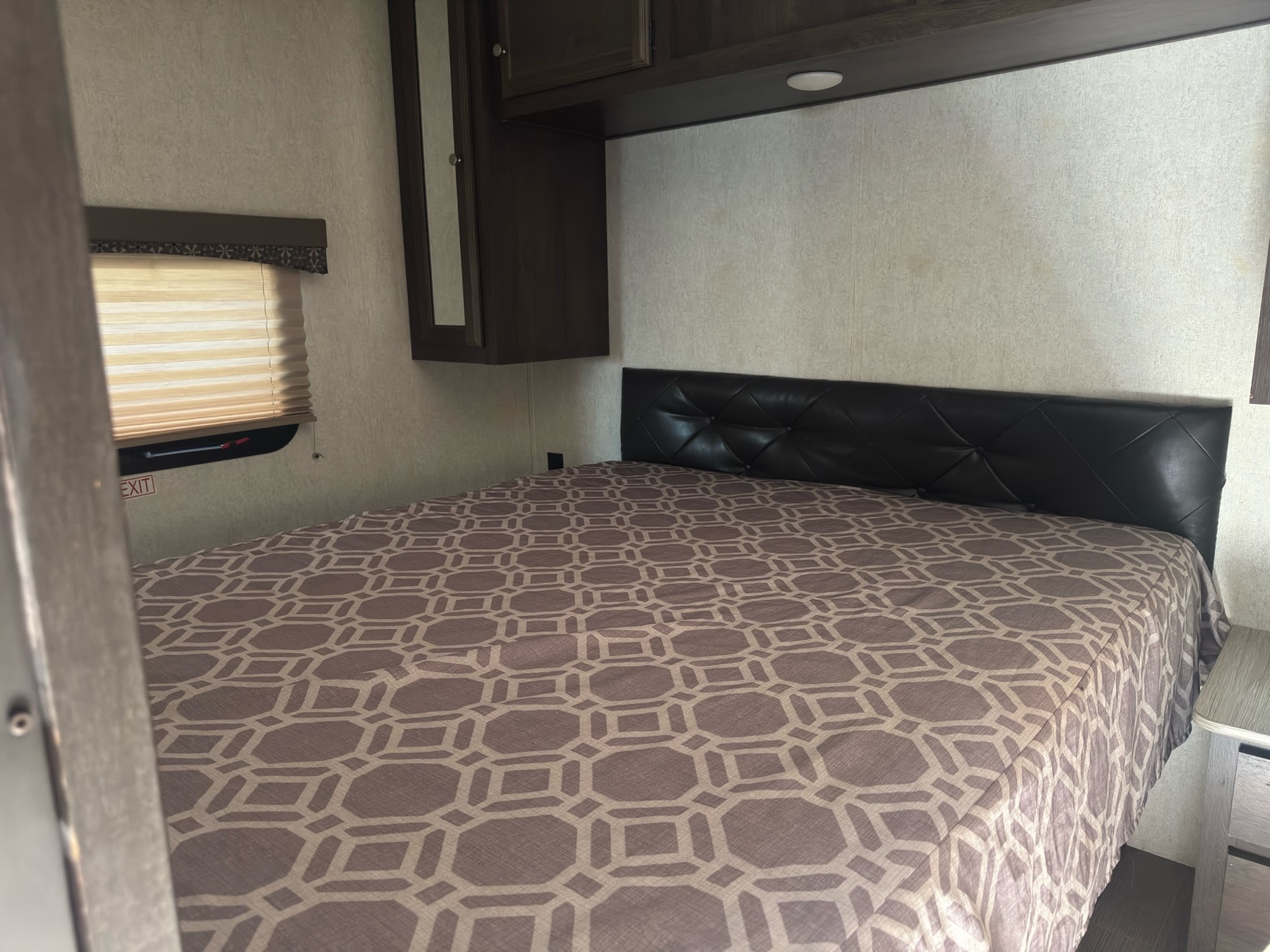 RV rental bedroom with a geometric bedspread, padded black headboard, window with blinds, and dark wood overhead cabinets.. Starcraft GPS 2018