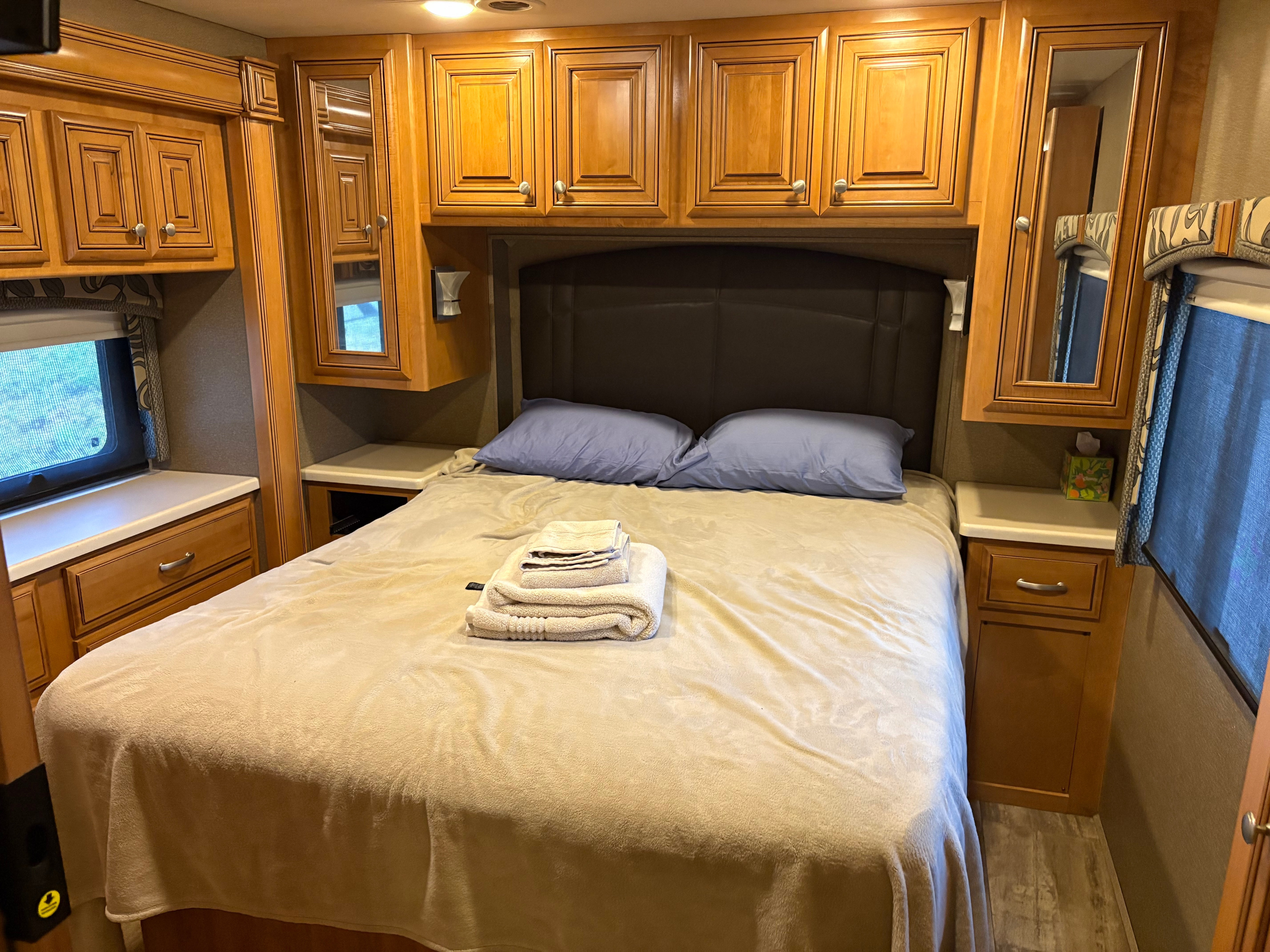 Inside an RV rental: a comfortable bedroom with a queen bed, overhead wood cabinets, and side windows.. Thor Palazzo 2016