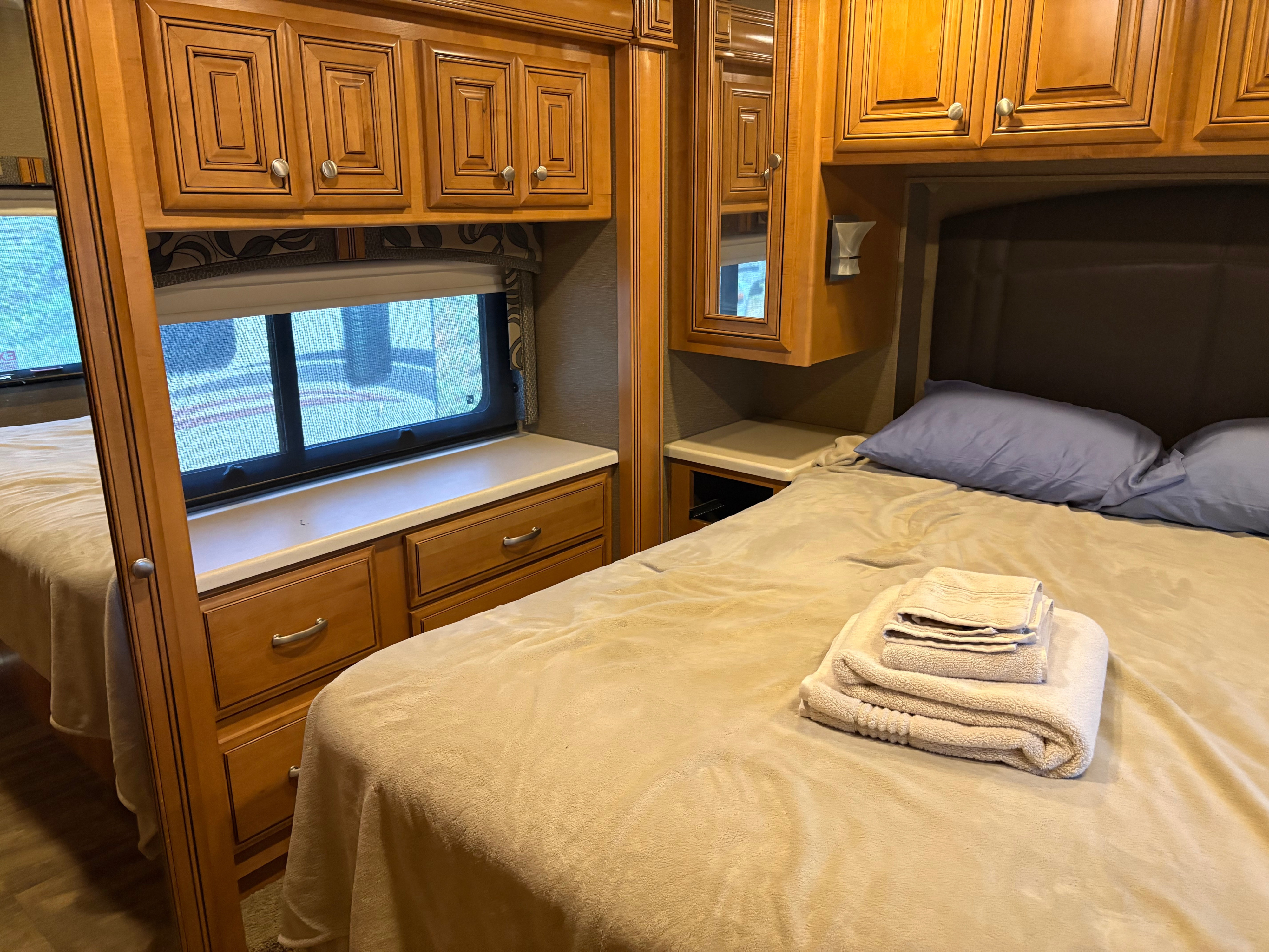Cozy RV rental bedroom with two beds, warm wood cabinetry, a window, and fresh towels for a comfortable journey.. Thor Palazzo 2016