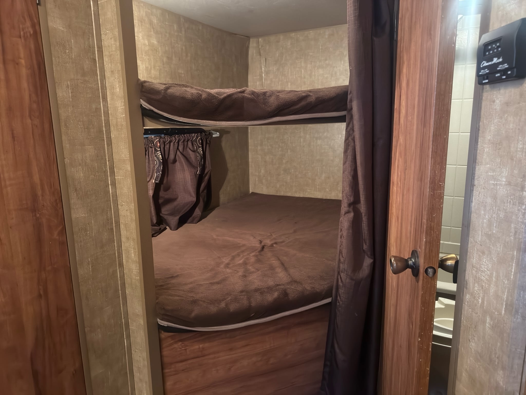 RV rental bunk beds with brown bedding and privacy curtains. A thermostat and glimpse of the bathroom are visible on the right.. KZ C291IKS 2015