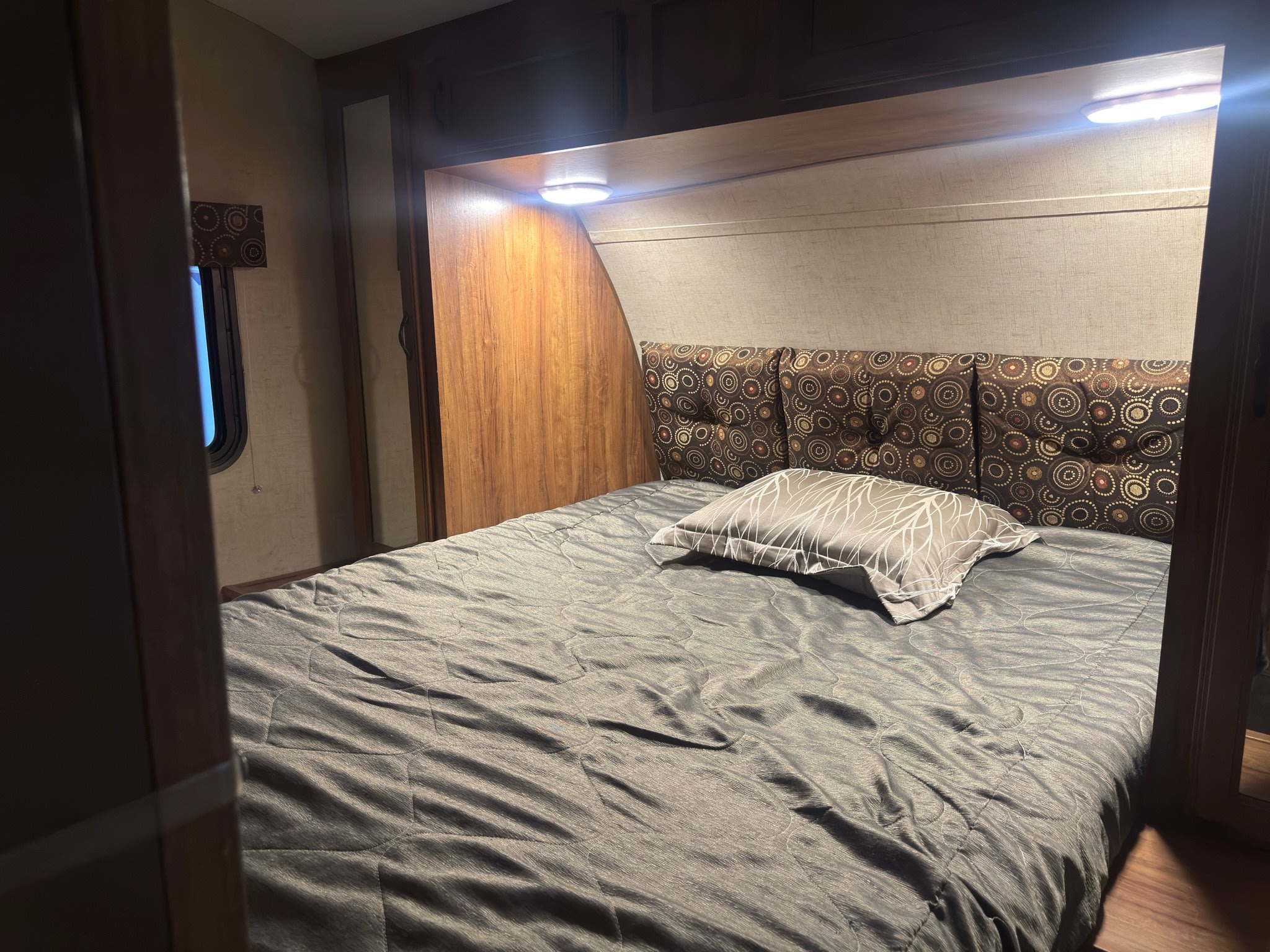 Cozy RV rental bedroom features a patterned headboard, grey comforter, and overhead lighting for comfortable travel.. KZ C250BHS 2015