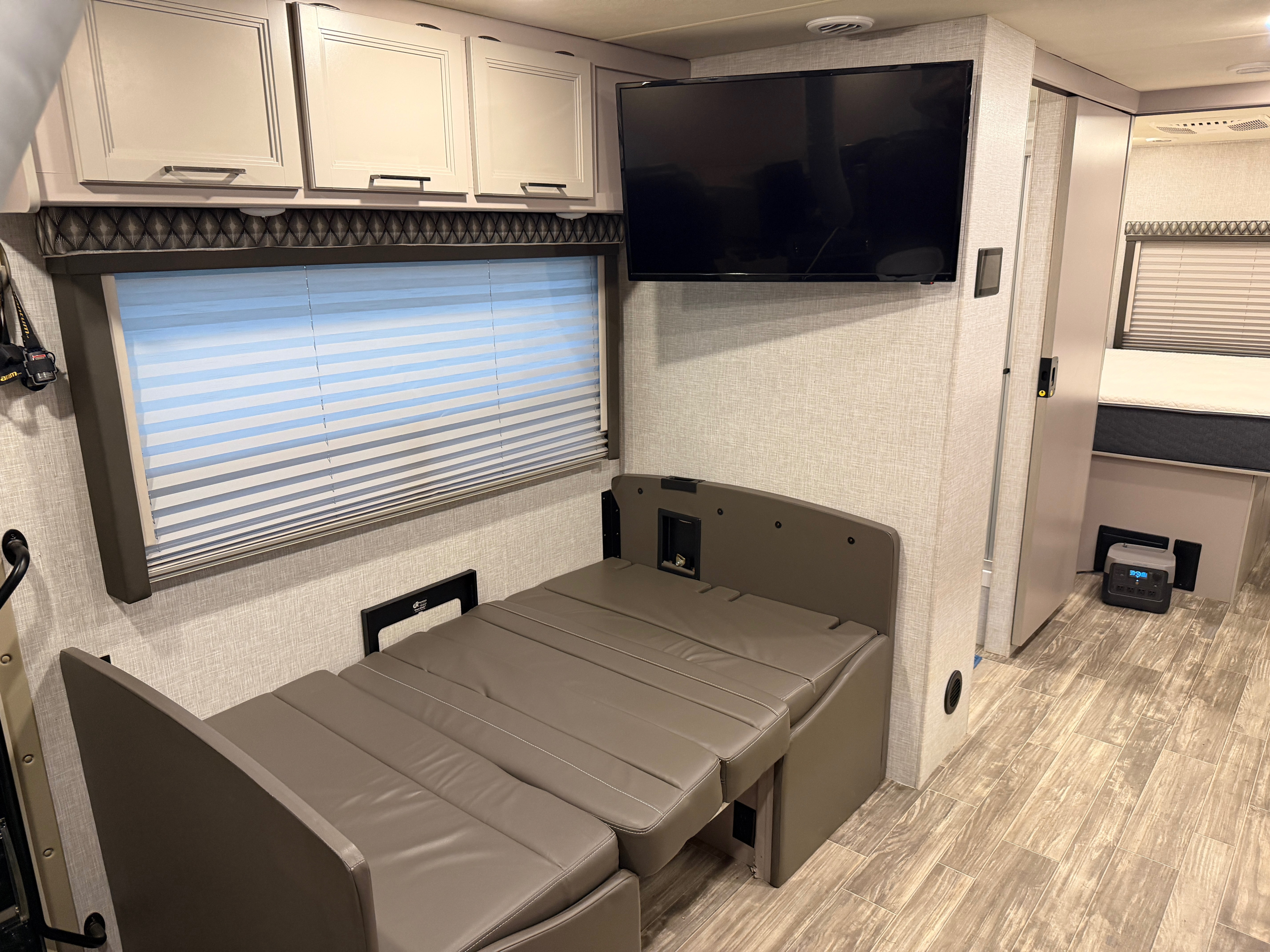 Modern RV rental interior featuring a taupe convertible sofa/bed, wall-mounted TV, light overhead cabinets, and wood-look floor.. Thor Hurricane 2022