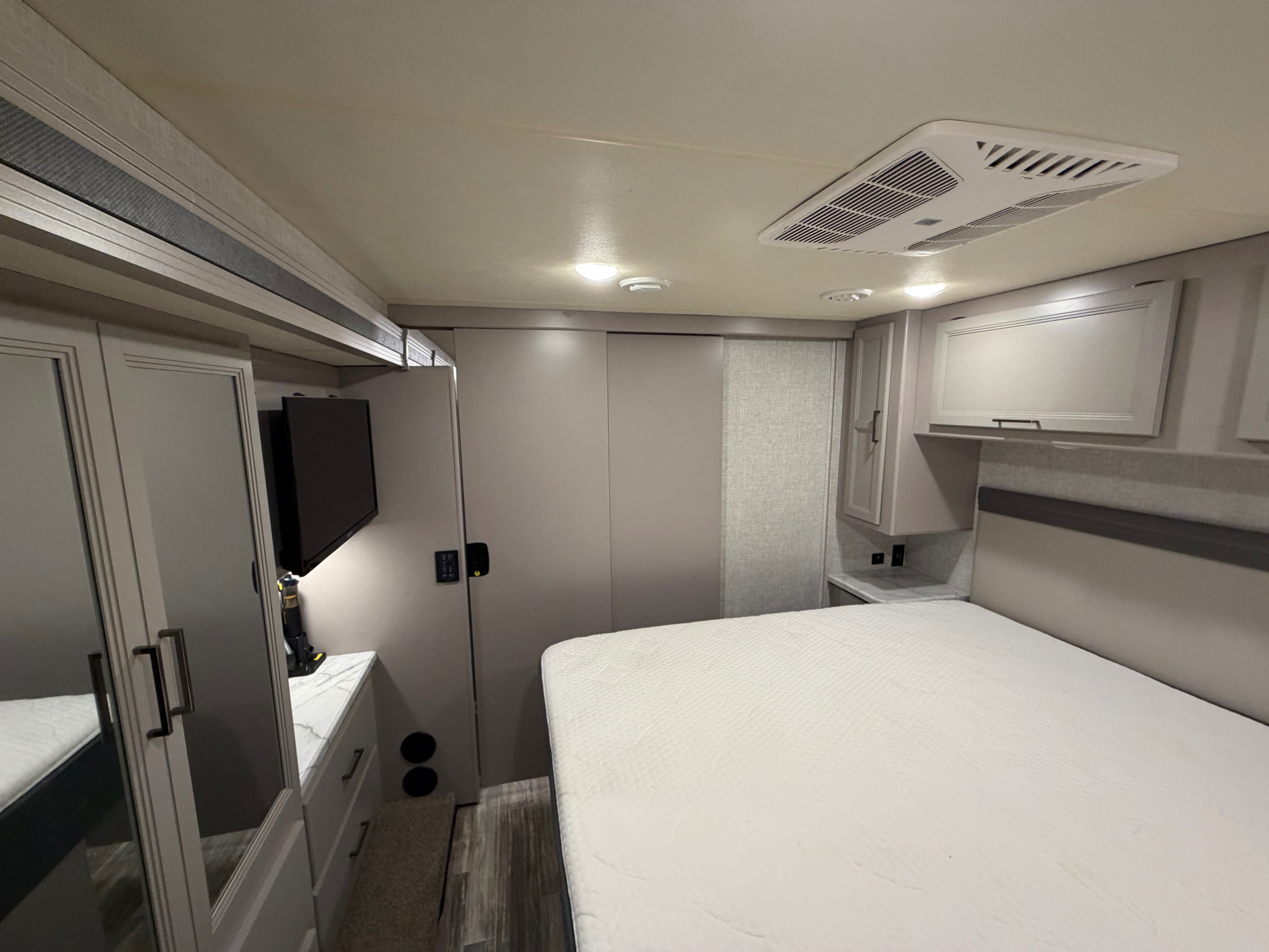 Light-toned RV rental bedroom: white bed, overhead AC, TV, and gray cabinets. Ideal for comfortable travels.. Thor Hurricane 2022