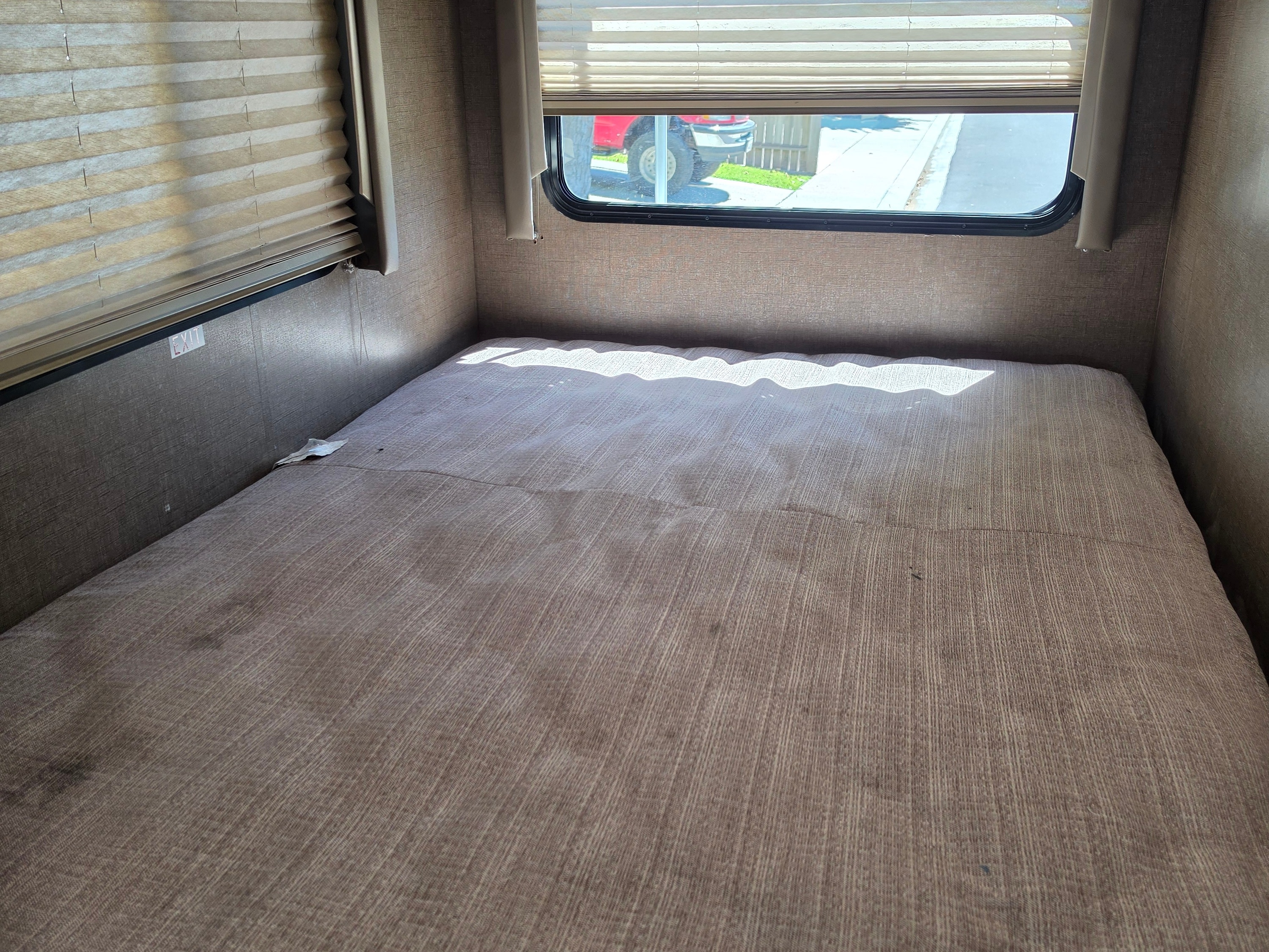 RV rental bedroom: a brown textured bed under a window showing outside, with an EXIT sign on the wall. Sunlight patterns the bedding.. Other Other 2020