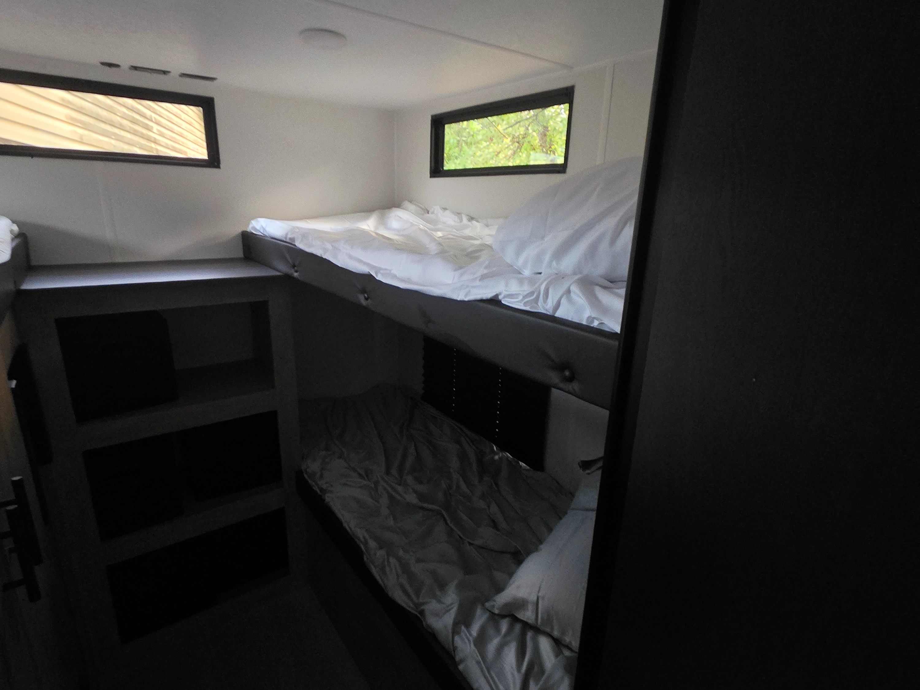 Inside an RV rental: bright bunk beds with white bedding, windows offering views of trees. Built-in shelving unit is on the left.. Forest River 29TJ 2027