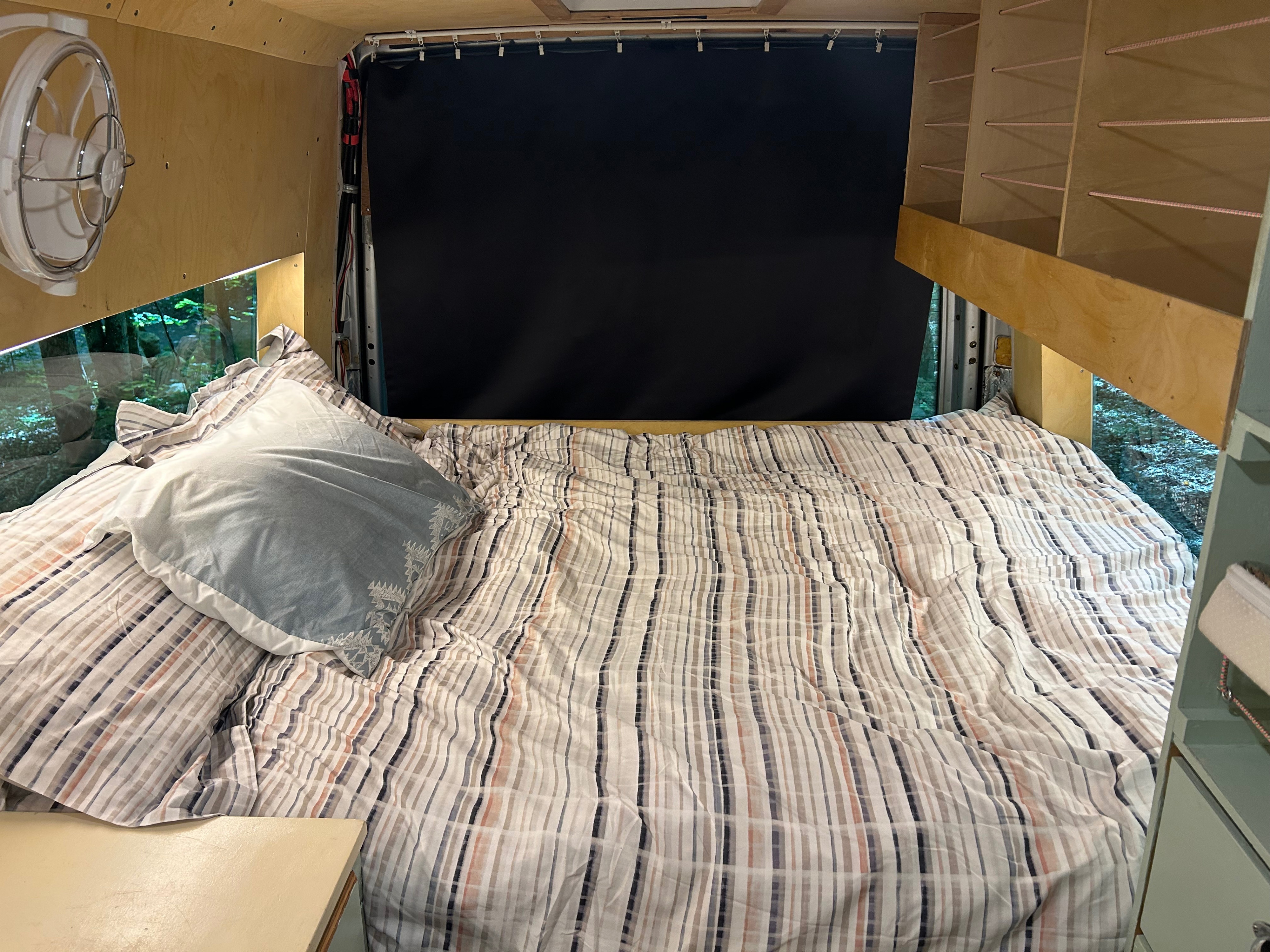 RV rental's cozy bed with striped duvet, window forest view, wall fan, and built-in storage. Ideal for road trips.. Dodge Sprinter 2005