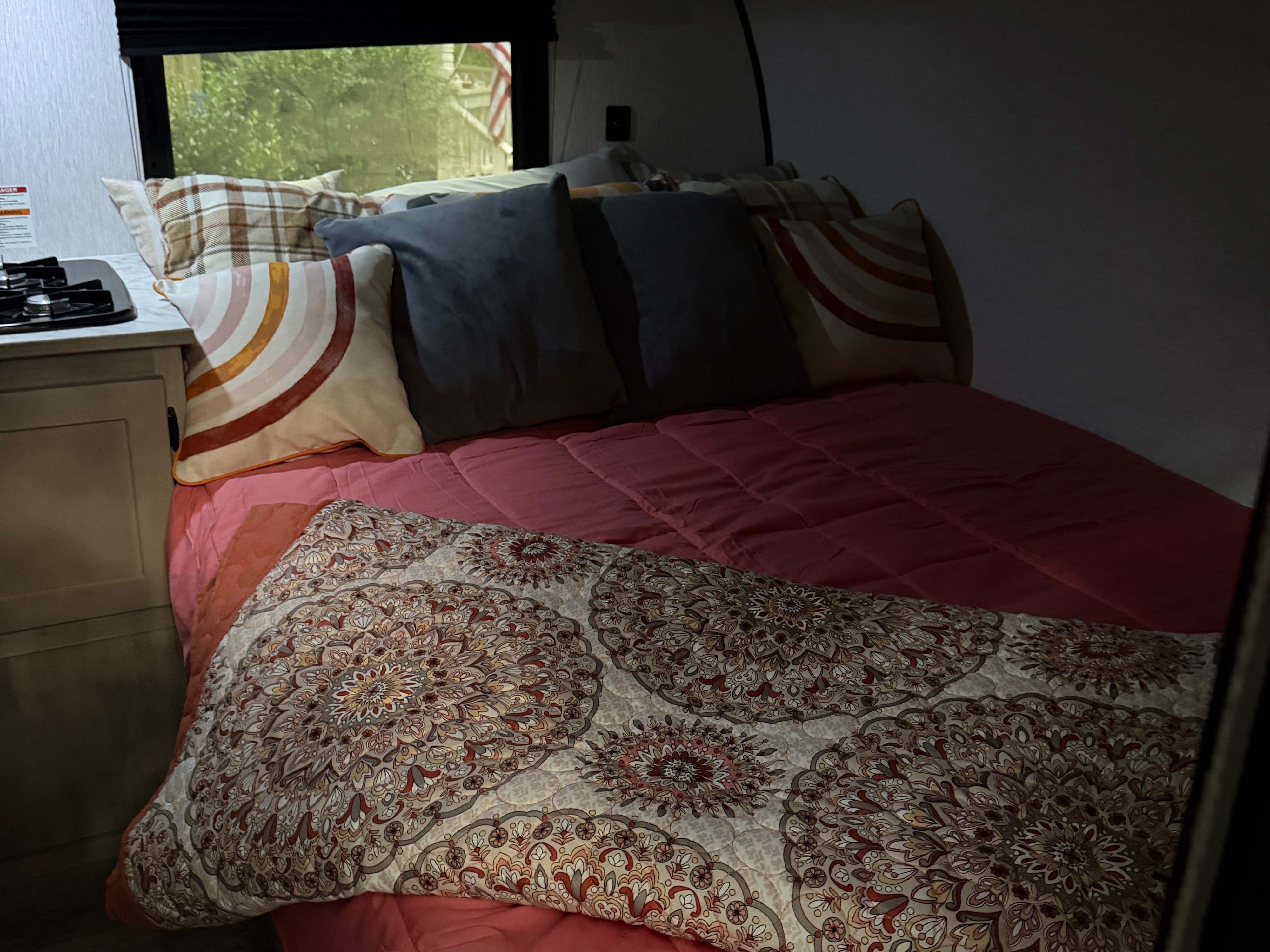 Cozy rv rental bed with colorful pillows, pink comforter, and patterned blanket. A compact stove and window view complete the interior.. Sunlite 13BHLTD 2027