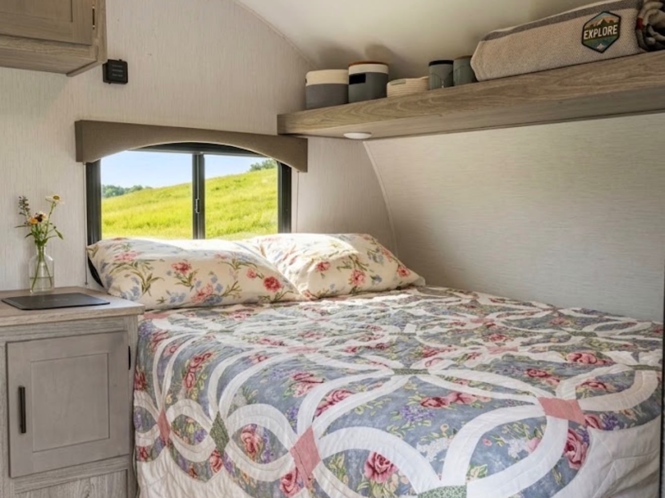 A cozy RV rental bedroom features a floral bed, window with a lush green view, and practical overhead shelving.. Sunset Park RV Sun Lite LTD 2024
