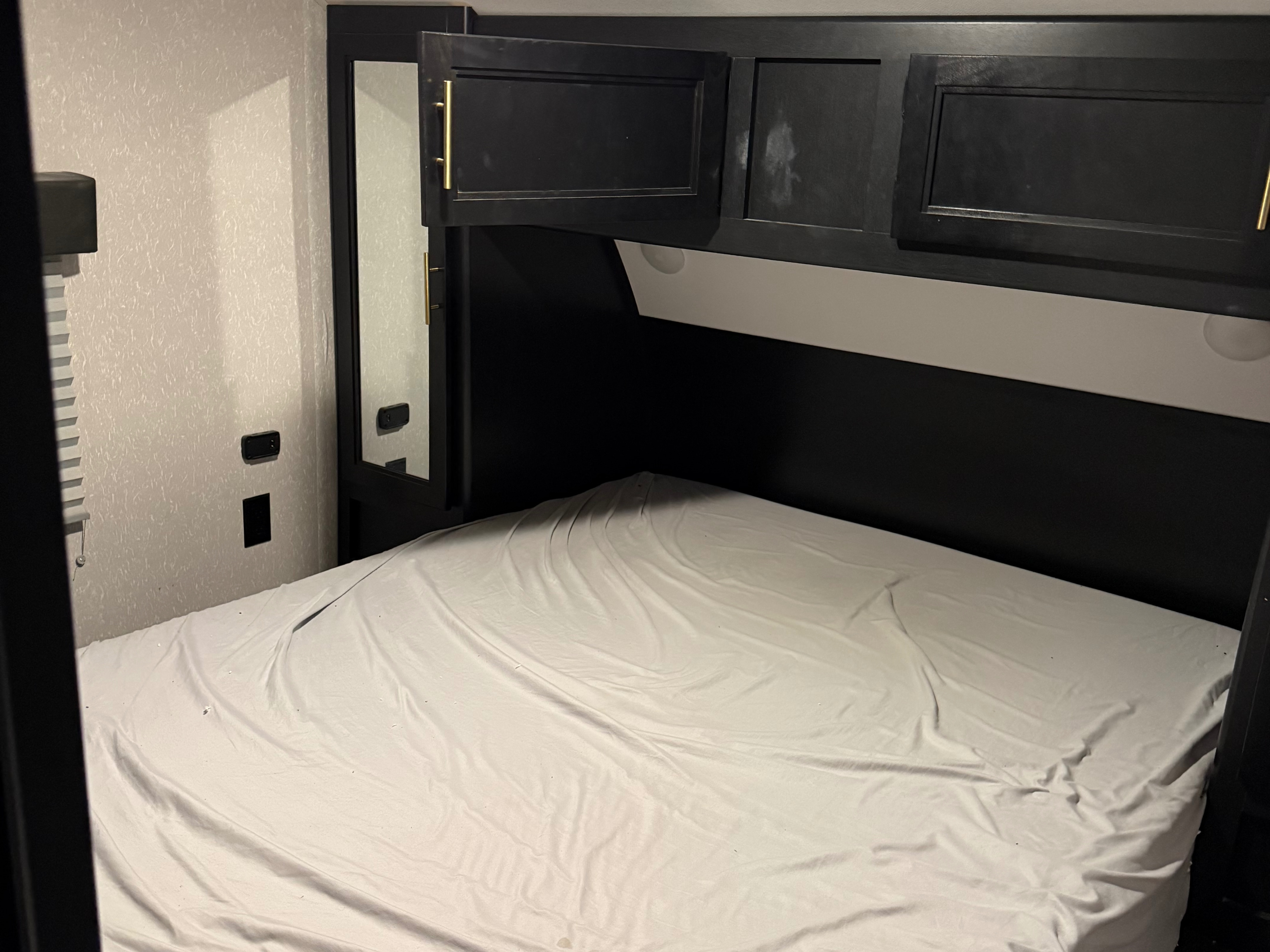 Inside an RV rental bedroom showing a bed with white sheets, overhead black cabinets, and a mirror.. Forest River Cherokee 2023