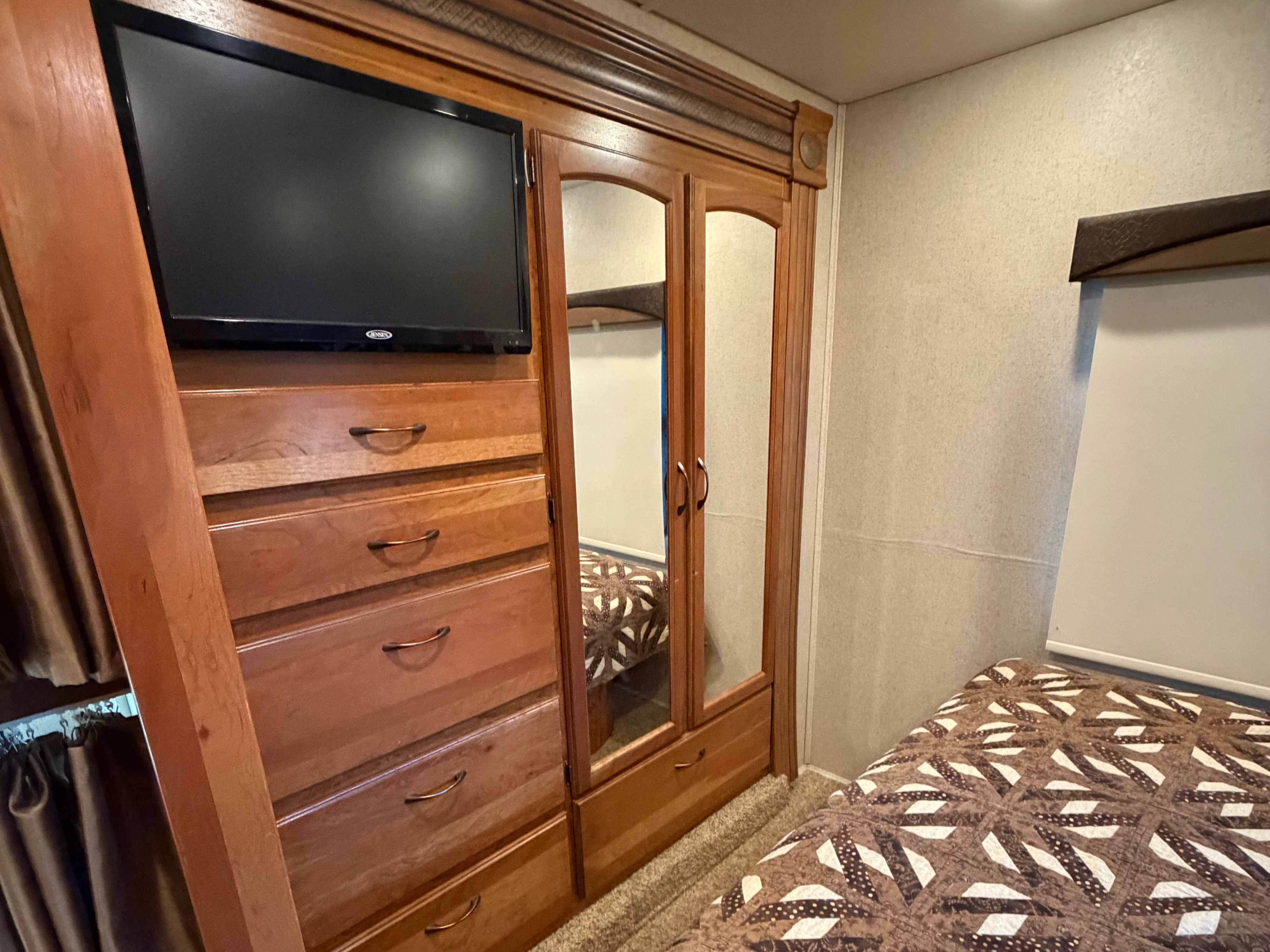 Compact RV rental bedroom featuring a TV, wooden dresser, mirrored closet, and a patterned bed quilt.. Jayco Greyhawk 2016