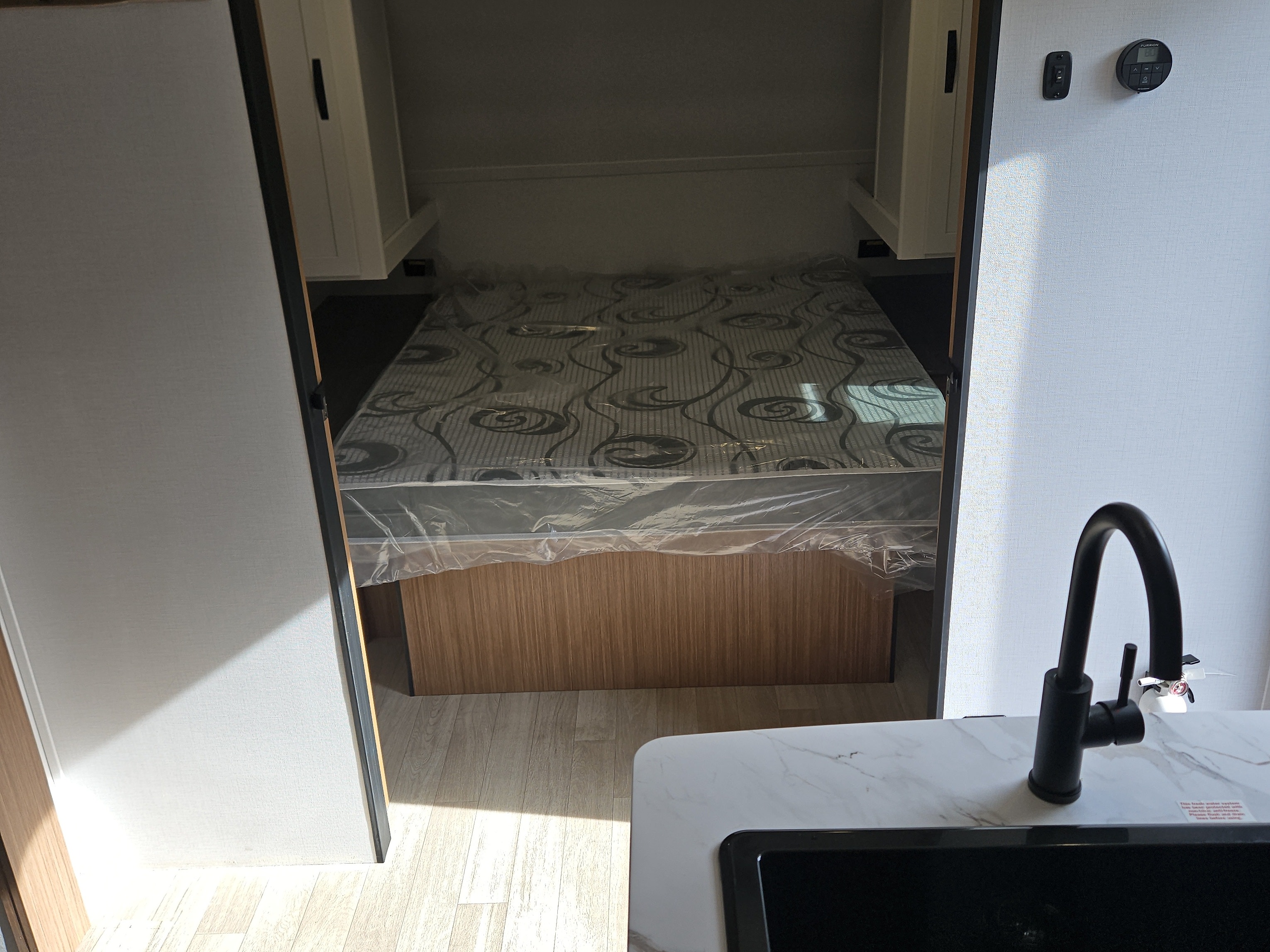 RV rental bedroom & kitchen view: new mattress with plastic, cabinets, a modern sink, and smart thermostat on light walls.. Keystone RV Passport Classic 2025