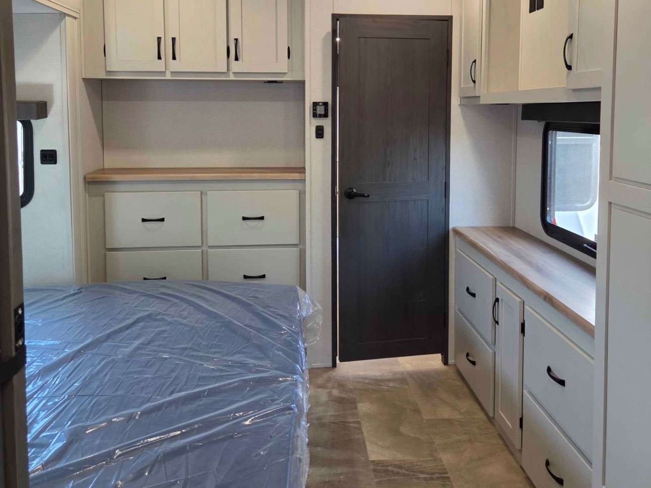 Modern RV rental bedroom with light grey cabinets, wood counters, a covered bed, and a dark door, showcasing a clean interior.. Coachmen Chaparral 2025