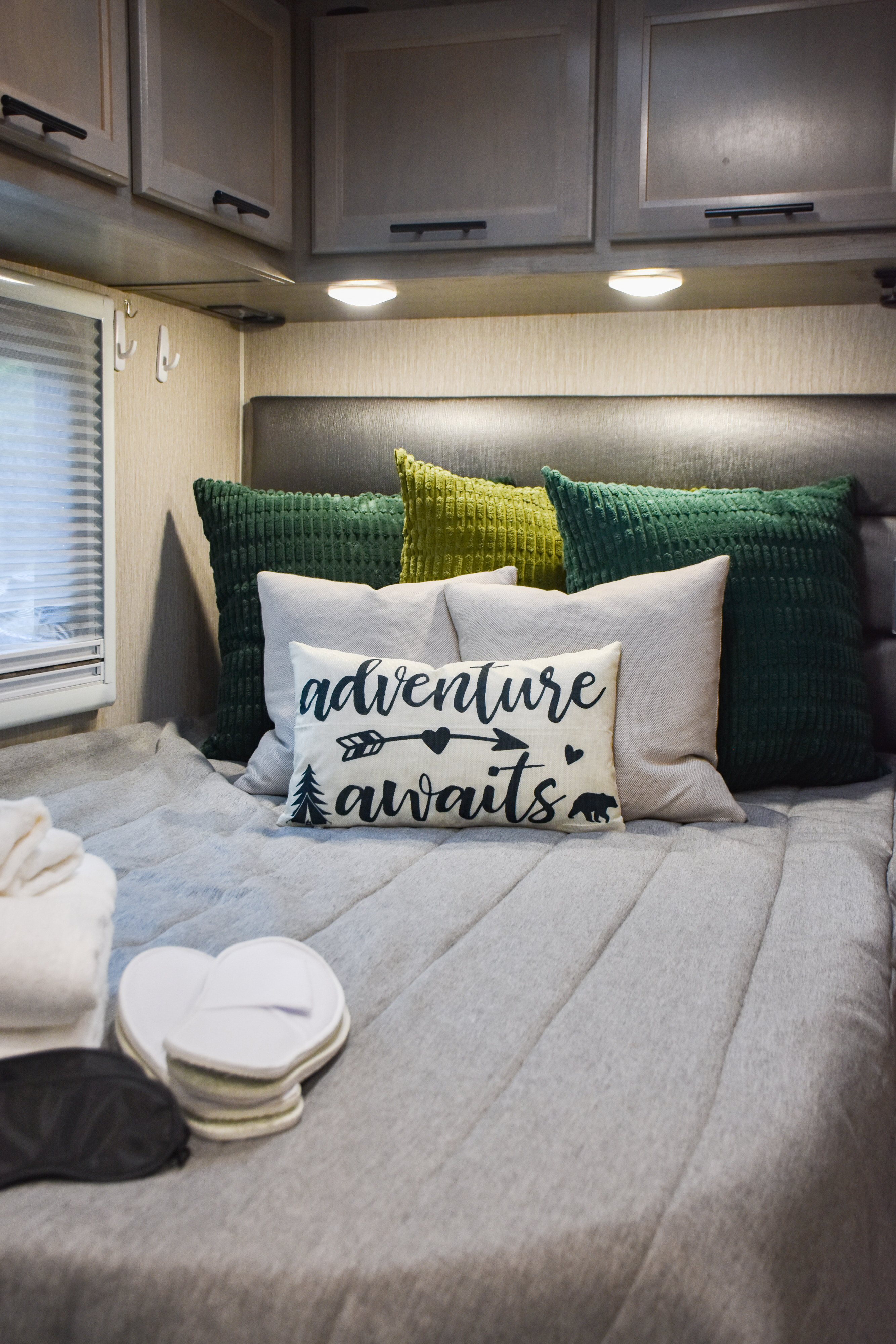 Cozy RV rental bedroom featuring a bed with plush green and grey pillows, an "adventure awaits" pillow, overhead cabinets, and a window.. Coachmen Prism 2022