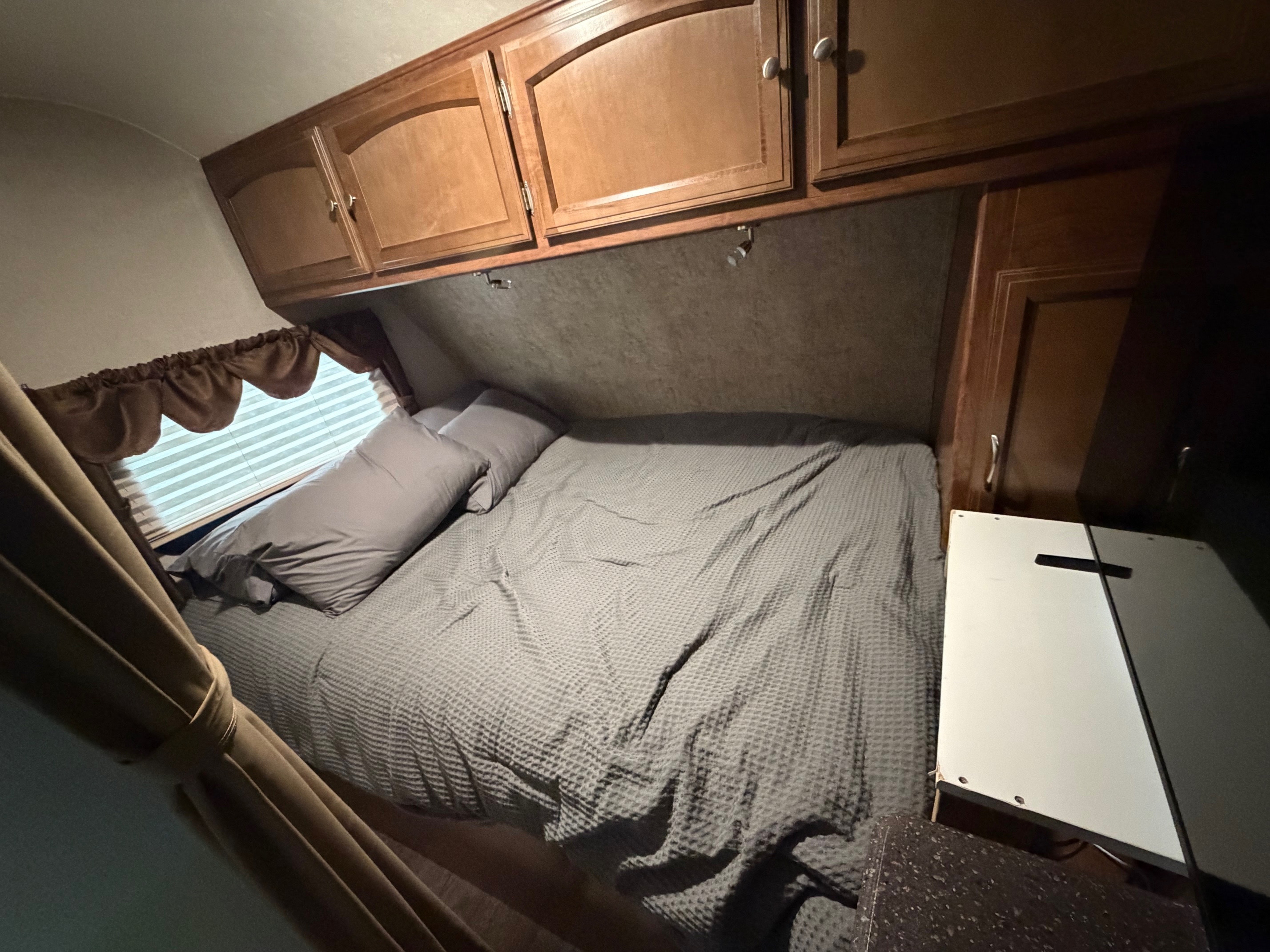 A compact RV rental bedroom with a bed, gray bedding, overhead wooden cabinets, a window with blinds, and curtains.. Coachmen Freedom Express 2017