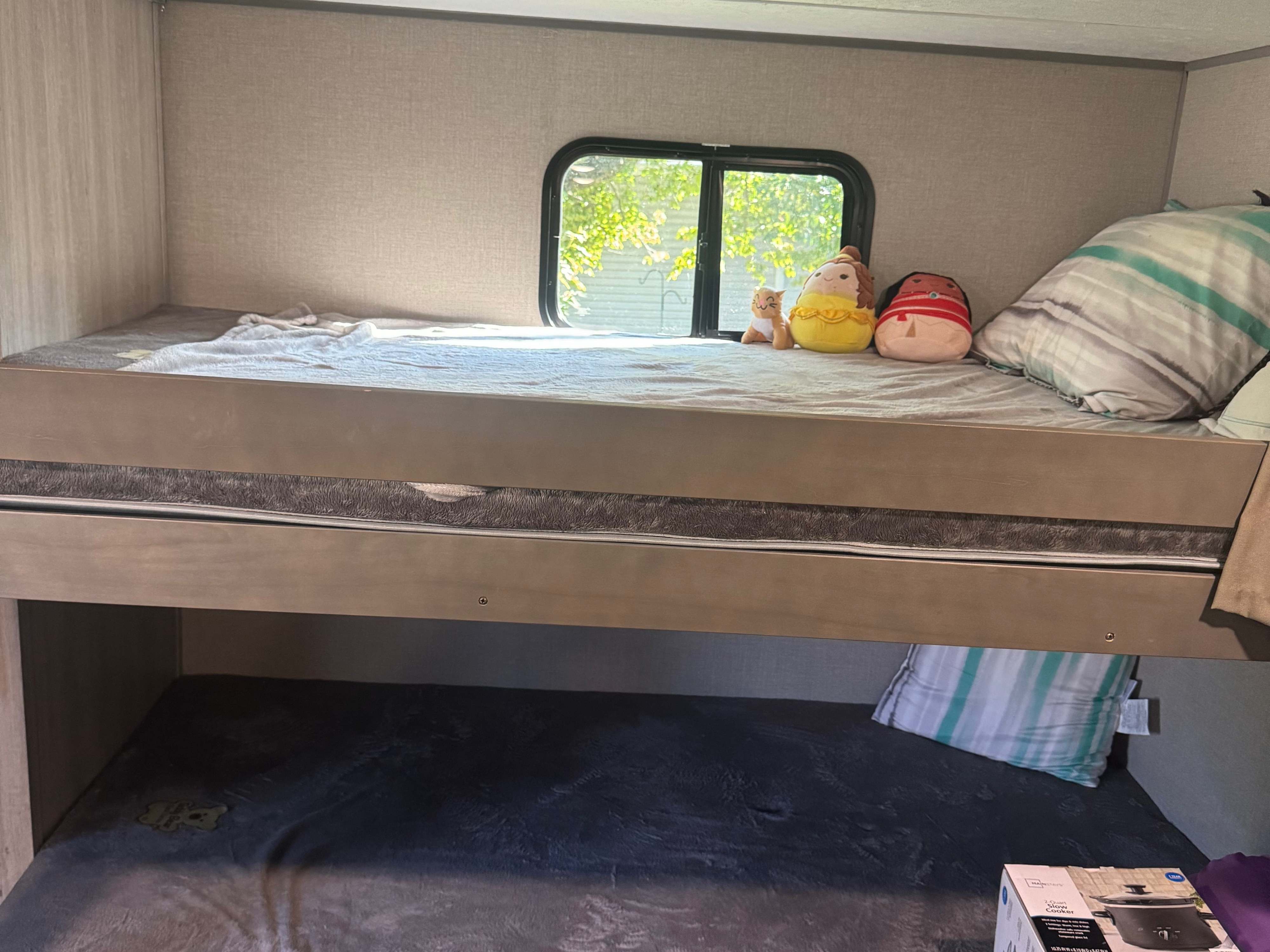 Inside an RV rental: cozy bunk beds. The top bunk has a window and plushies; the bottom has a blanket and slow cooker.. Coachmen Catalina summit series 8 2023