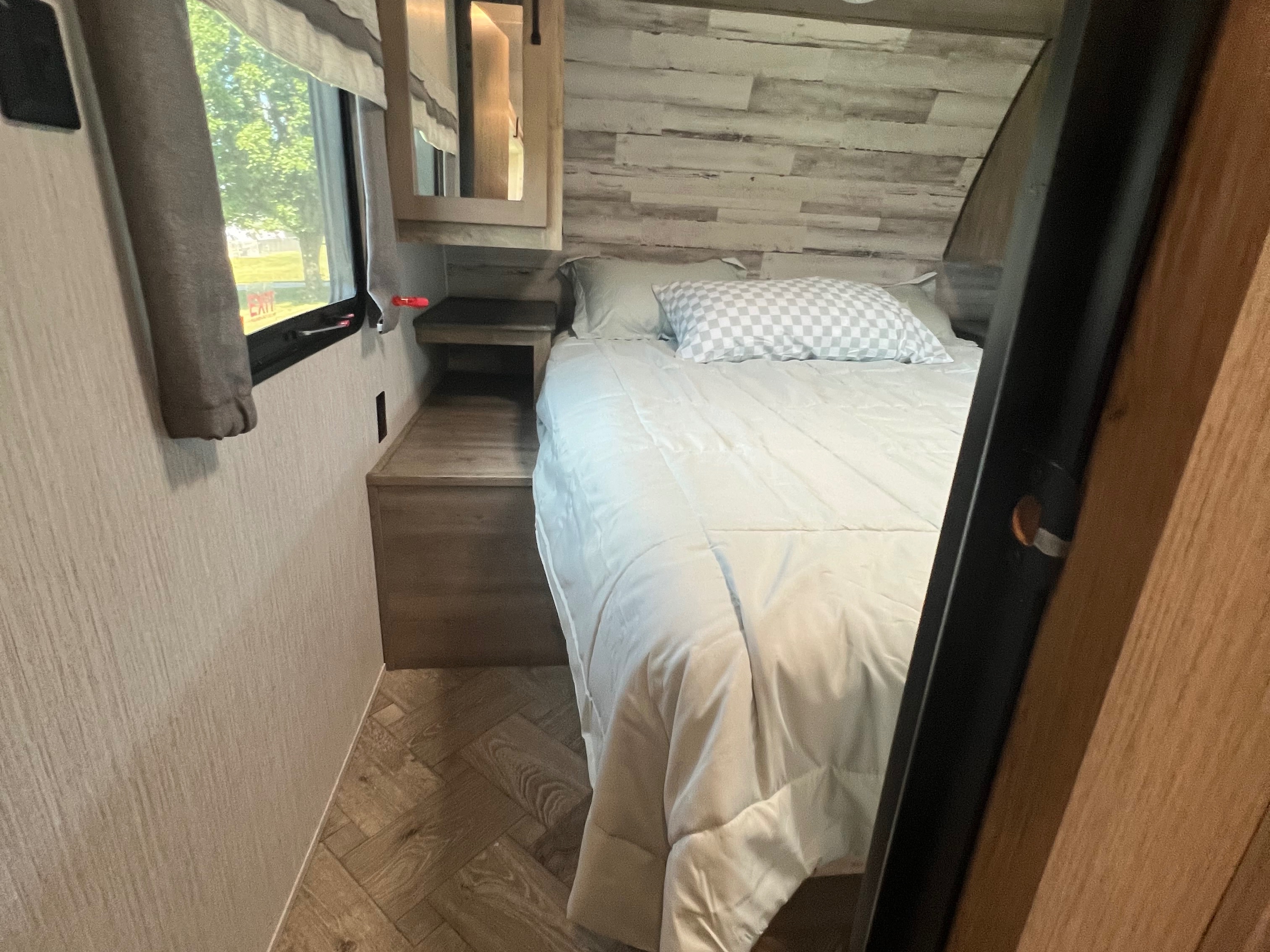 Cozy RV rental bedroom features a comfy bed, wooden nightstand, and window overlooking greenery, perfect for relaxing.. Forest River Palomino Puma 2021