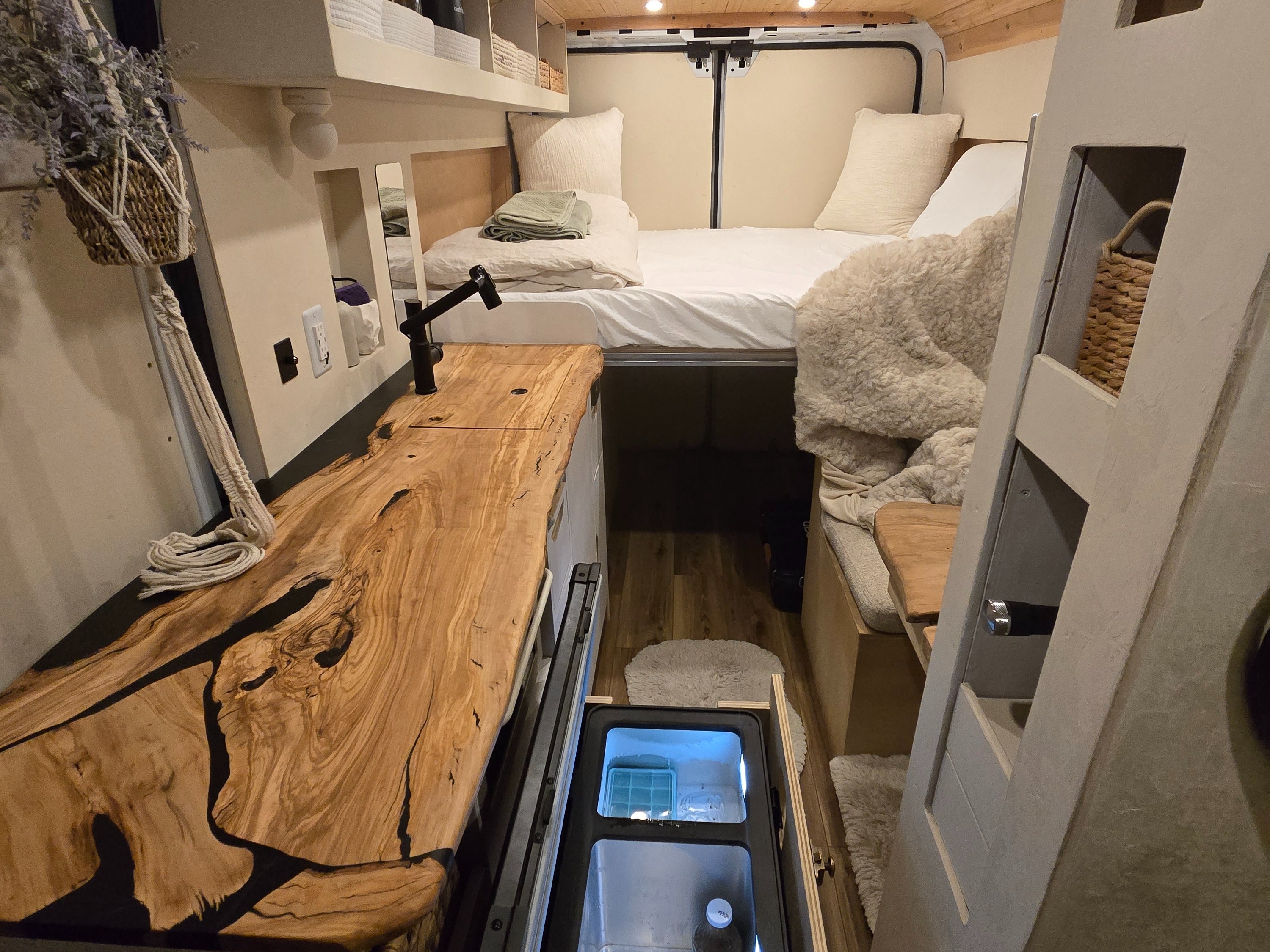 Cozy rv rental campervan interior with custom wood counter, pull-out fridge, bed, and a hanging macrame plant.. Dodge 1500 2023