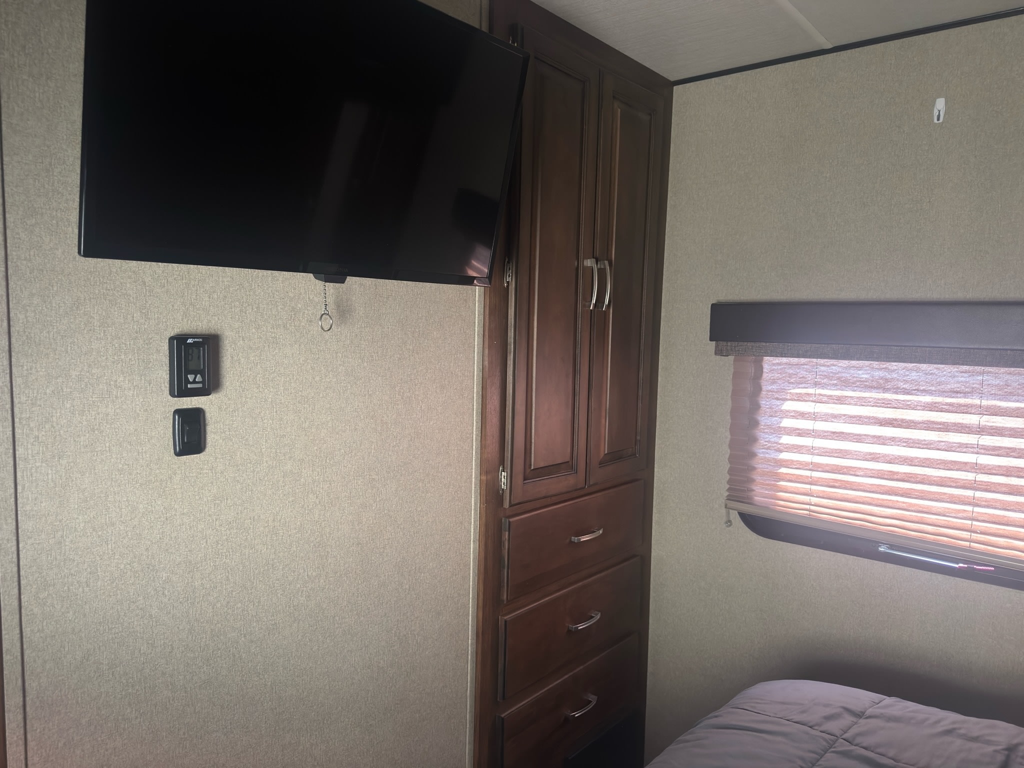 Interior of an RV rental bedroom showing a wall-mounted TV, dark wood cabinet, and a window with blinds over a bed.. Grand Design Reflection 2019