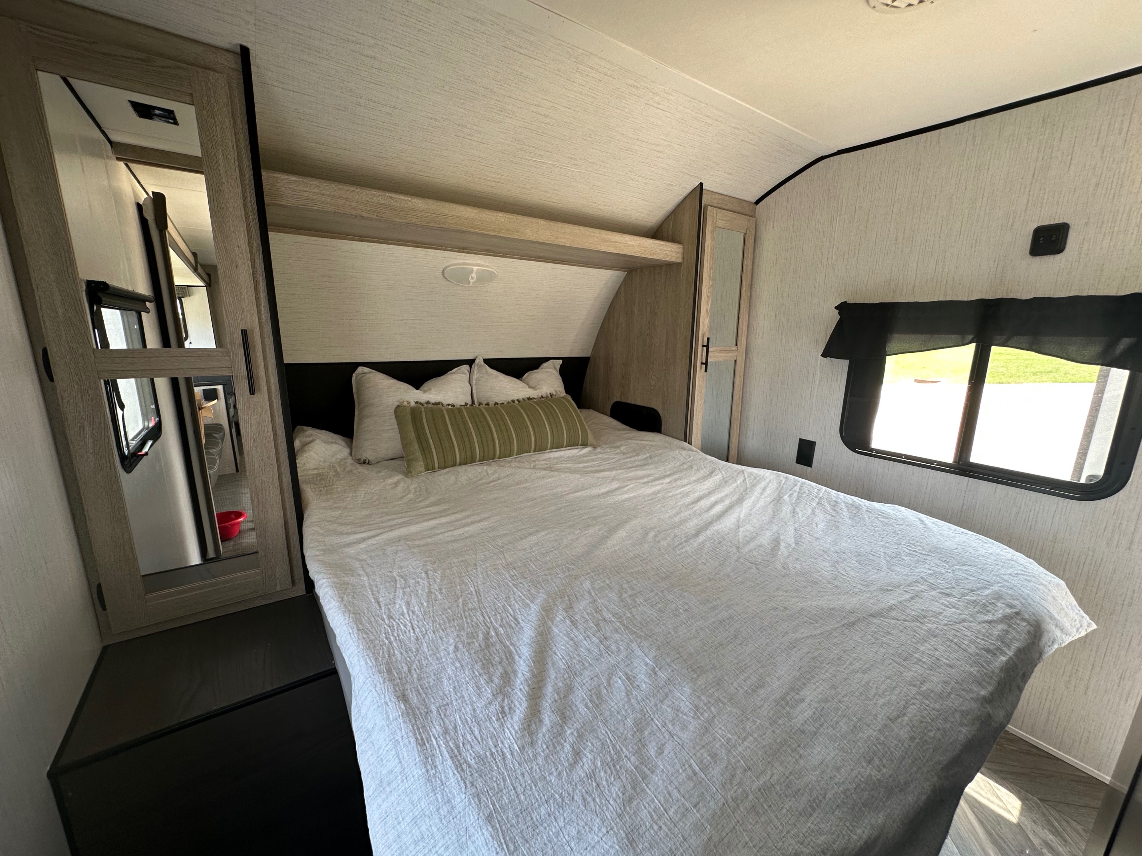 Inside an RV rental bedroom: a comfy bed with pillows, a window revealing nature, and wood-paneled storage.. Heartland RVs Prowler 2022