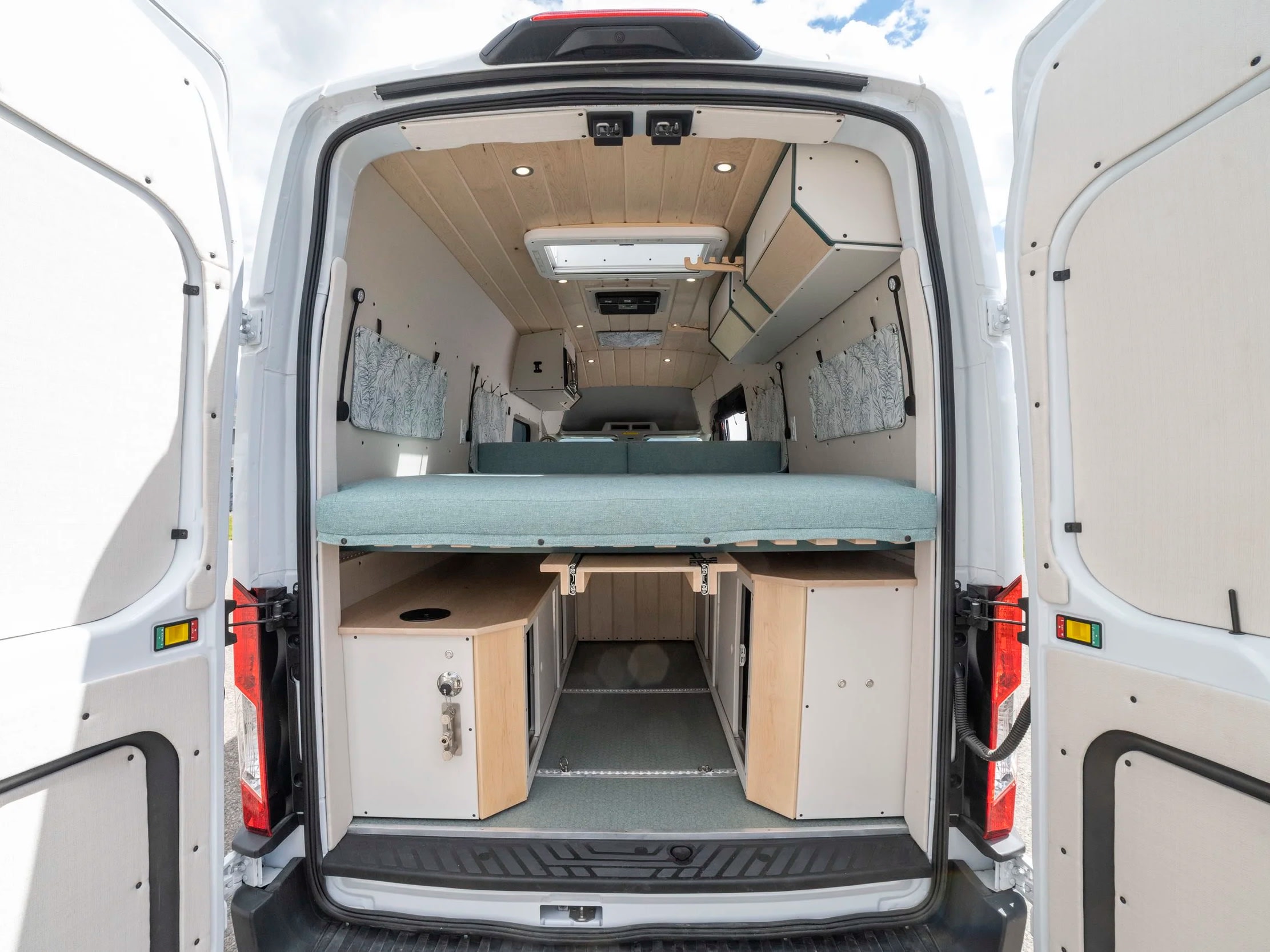 Open rear doors of a white RV rental reveal a custom camper van interior with a light blue bed, wood ceiling, and under-bed storage.. Ford Transit 2023
