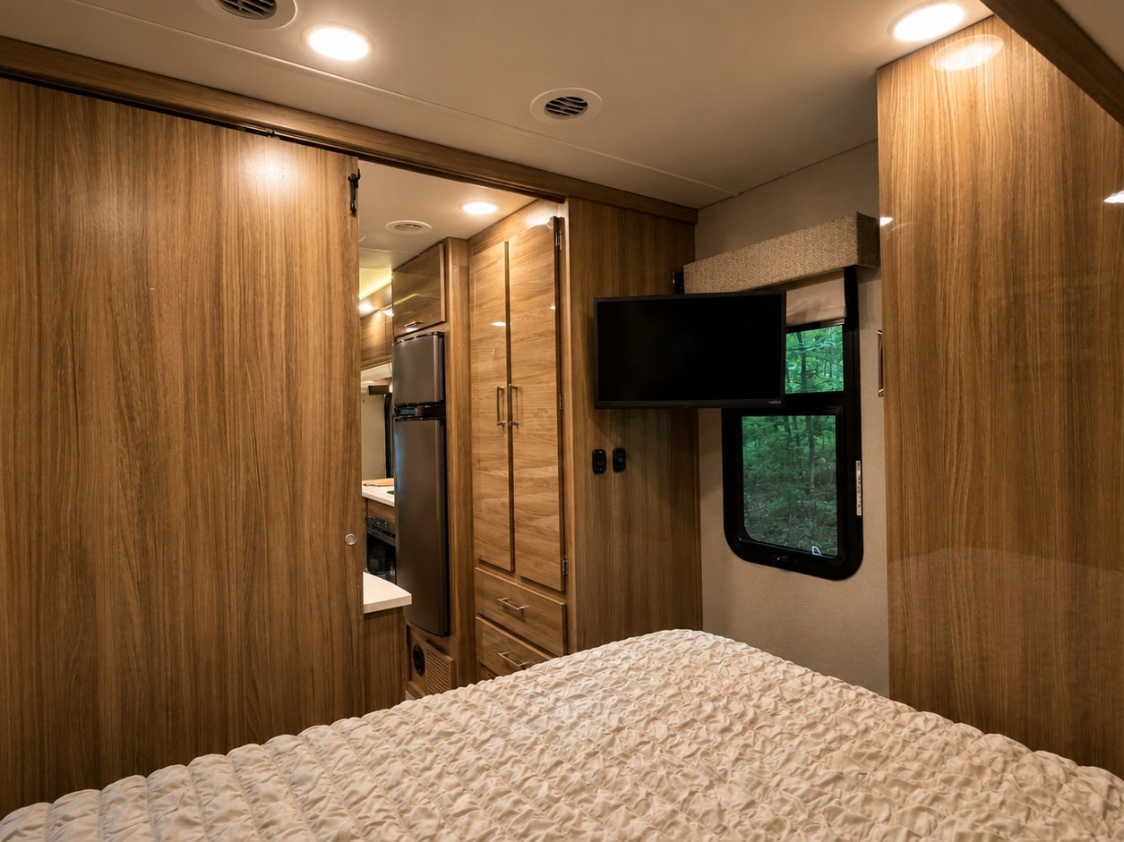 Stylish rv rental interior: bedroom with TV, window view, and sliding wood door to kitchen/living space.. Entegra Coach Qwest 2019