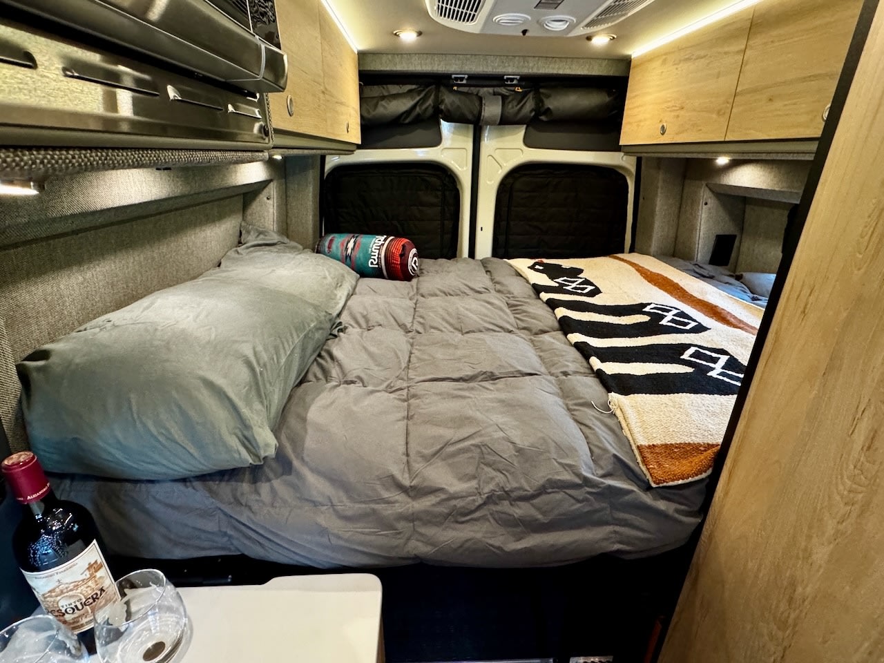 Cozy RV rental bedroom with a comfortable bed, stylish throw, and wine setup for a relaxing travel experience.. Storyteller Overland Storyteller Overland Class B 2025
