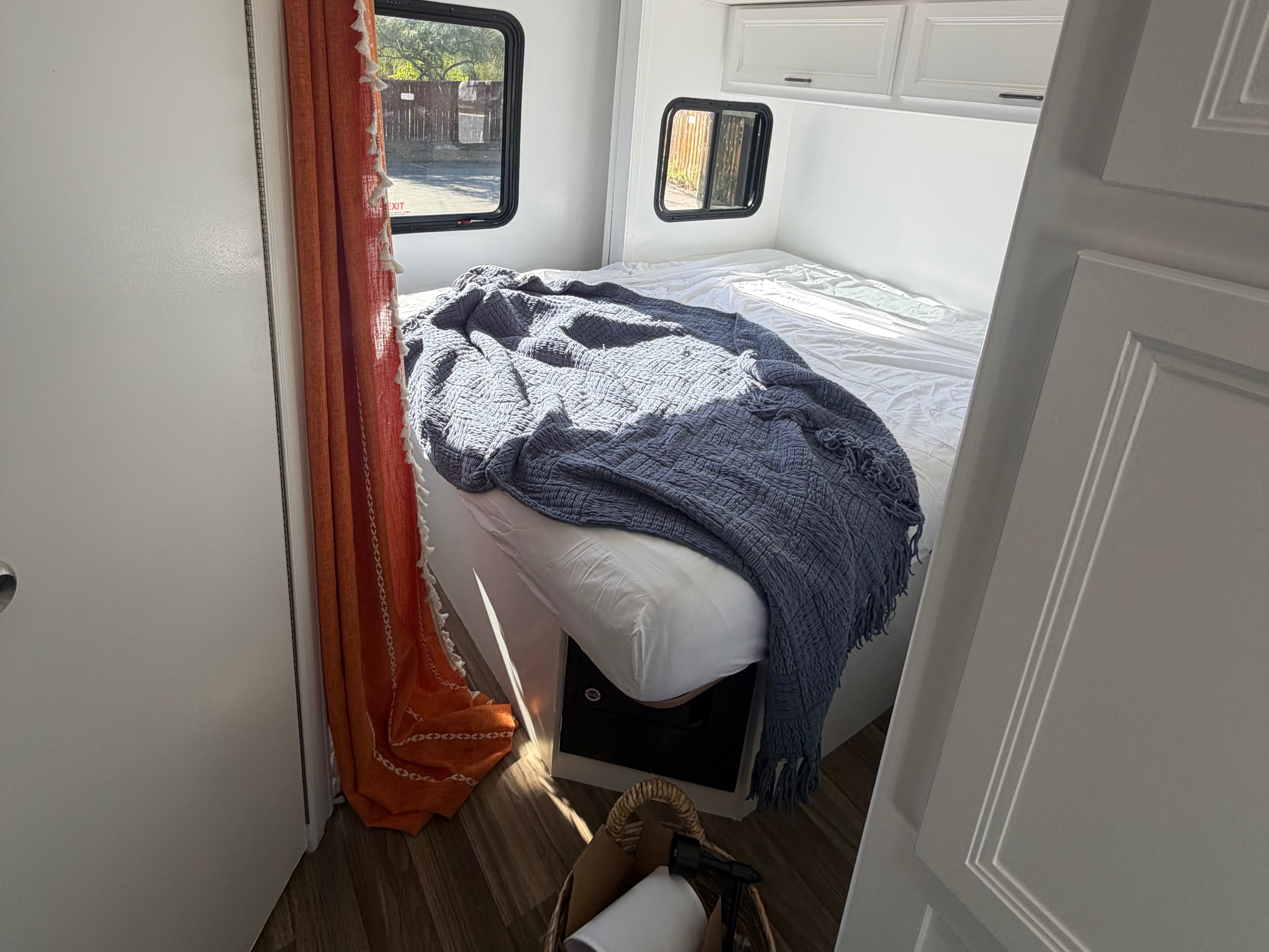 Bright RV rental bedroom: white bed, blue blanket, orange curtain, windows, and overhead cabinets, creating a cozy space.. Thor Quantum 2018