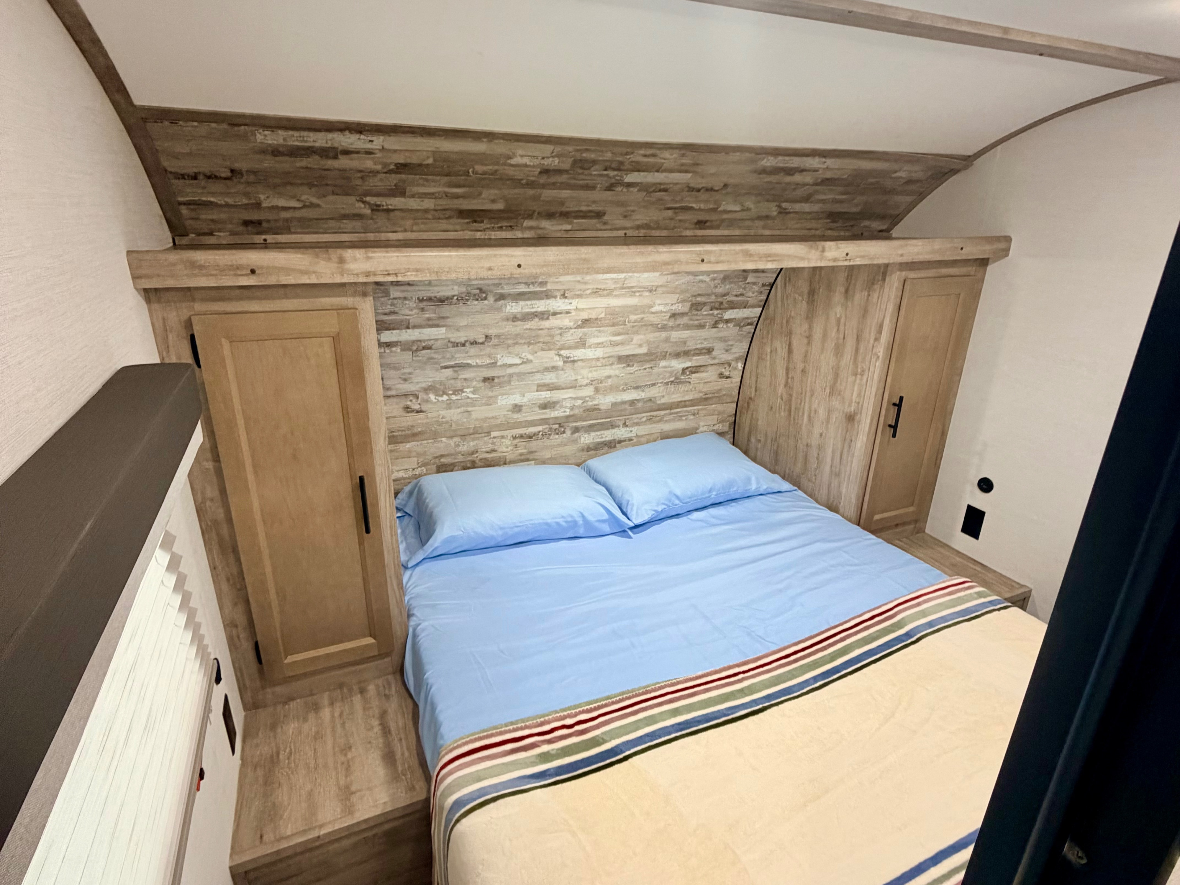 Inside a comfortable RV rental: a cozy bedroom features a bed with blue sheets, wood cabinets, and an accent wall.. Gulf Stream Enlighten SE 2025