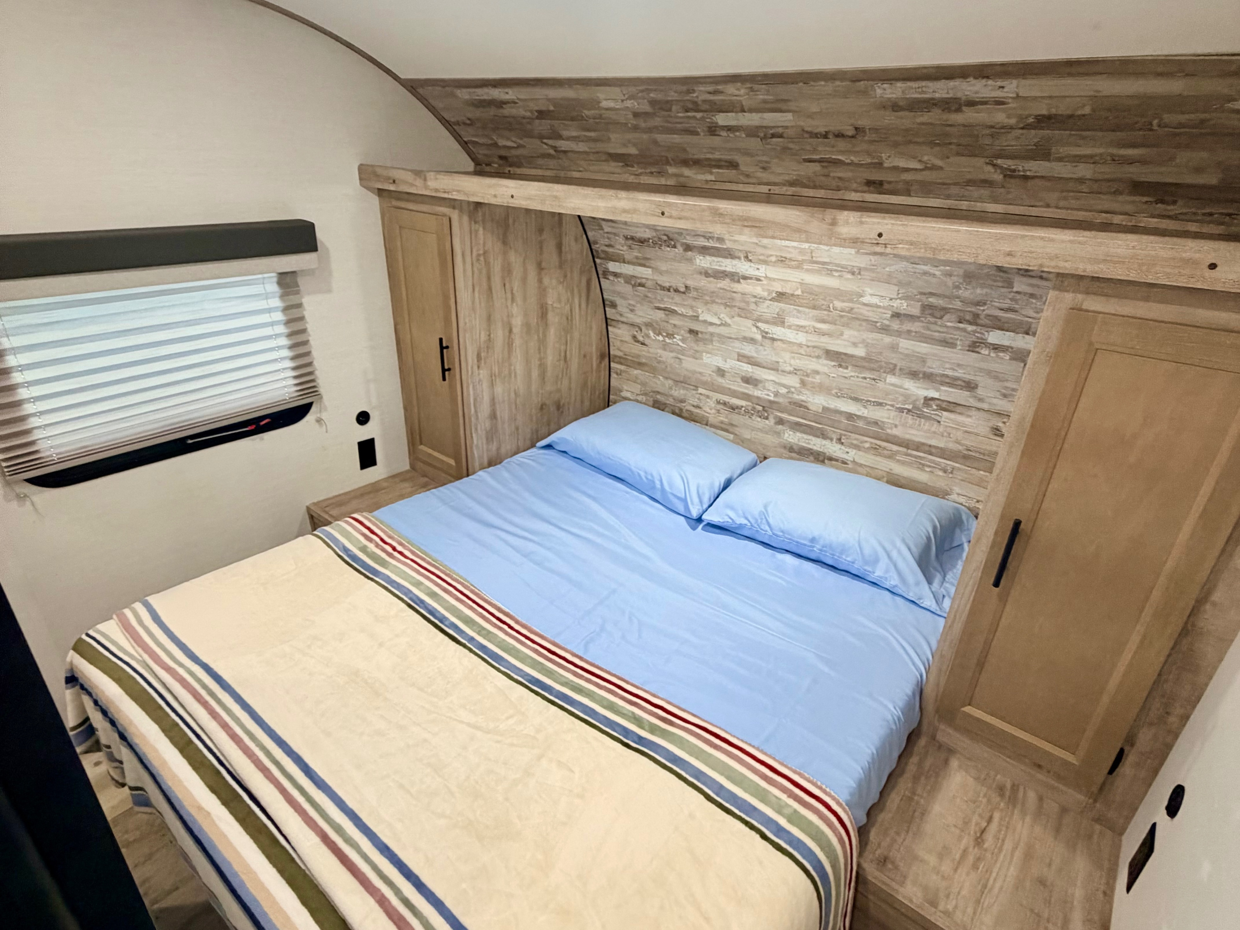 RV rental bedroom with a cozy bed, light blue sheets, striped blanket, and built-in wood-look storage.. Gulf Stream Enlighten SE 2025