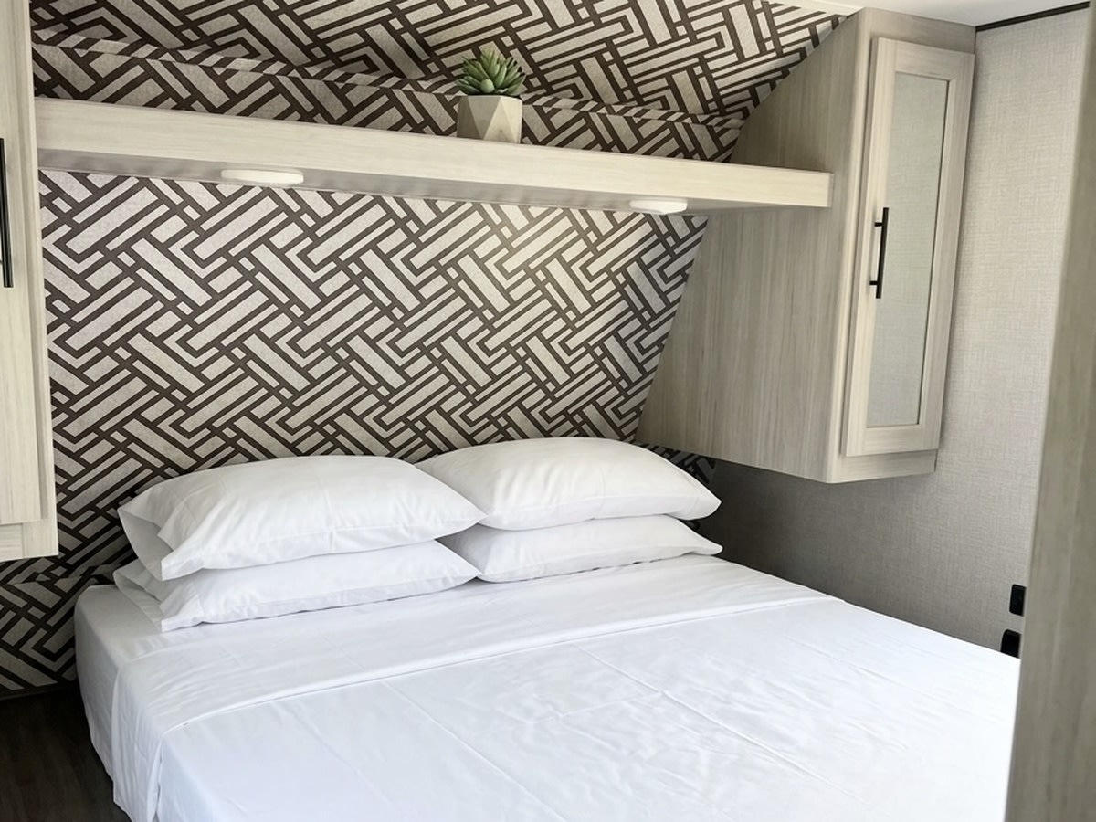 Modern RV rental bedroom showcasing a patterned headboard wall, white bedding, and space-saving overhead cabinets.. Crossroads RV Zinger 2024