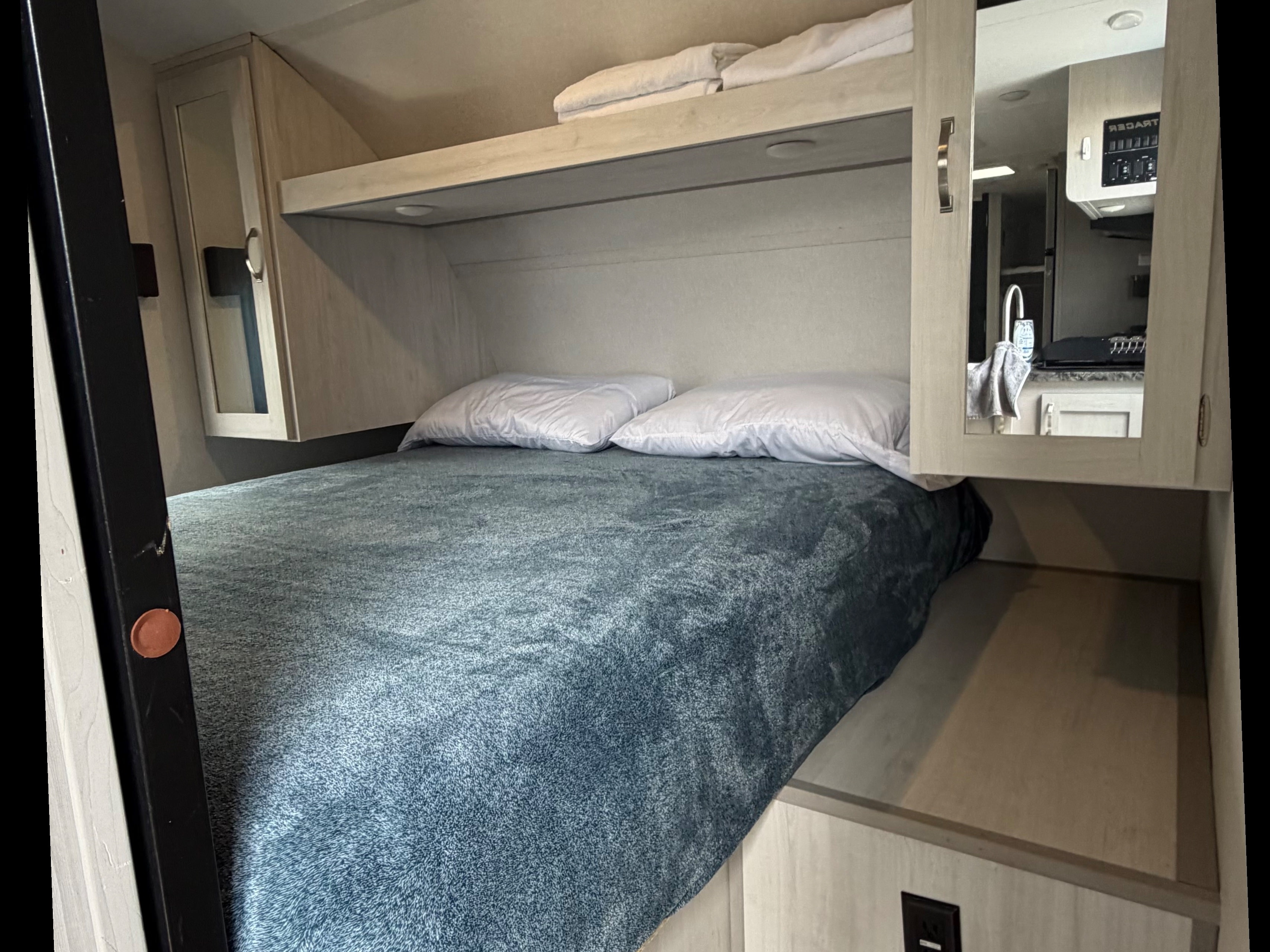 Cozy RV rental bedroom with bed, blue blanket, white pillows, and overhead storage cabinets. Mirrored doors reflect kitchen area.. Forest River Prime Time LE 2023