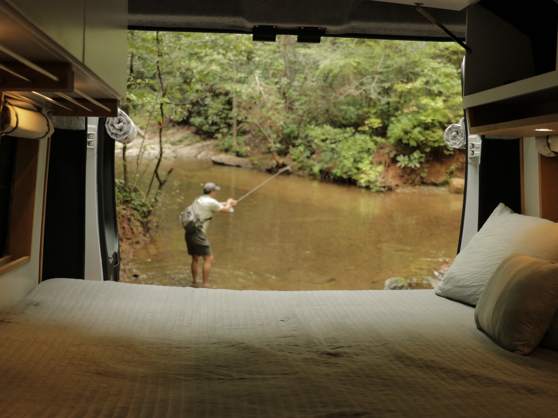 Experience serene fishing trips with our RV rental. A man fishes in a river, viewed from a cozy camper van bed.. Dodge Sprinter Van 2016