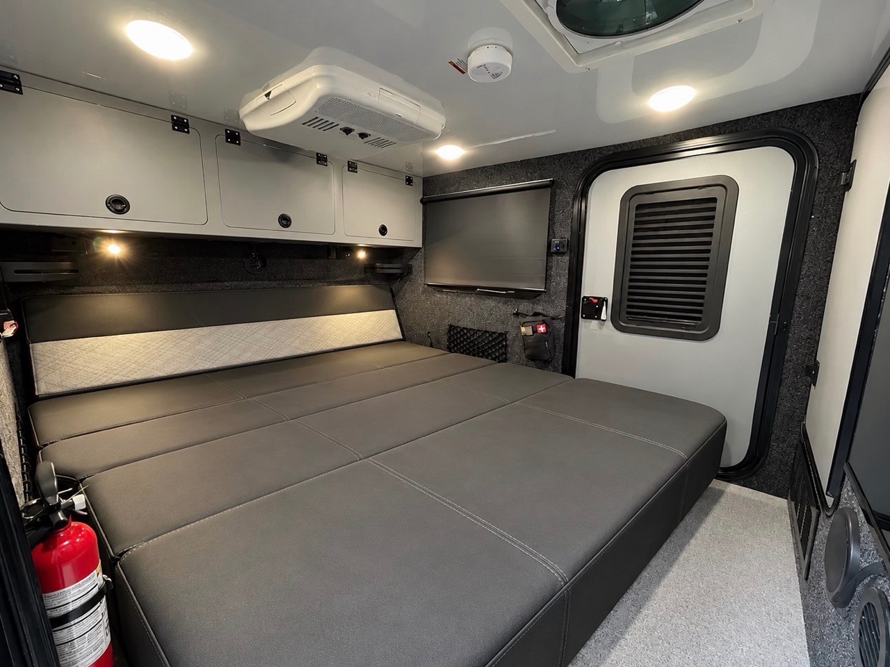 Inside a modern **RV rental**: A comfortable sleeping area with a large bed, overhead cabinets, AC unit, TV, and emergency exit.. Encore RV ROG 2026