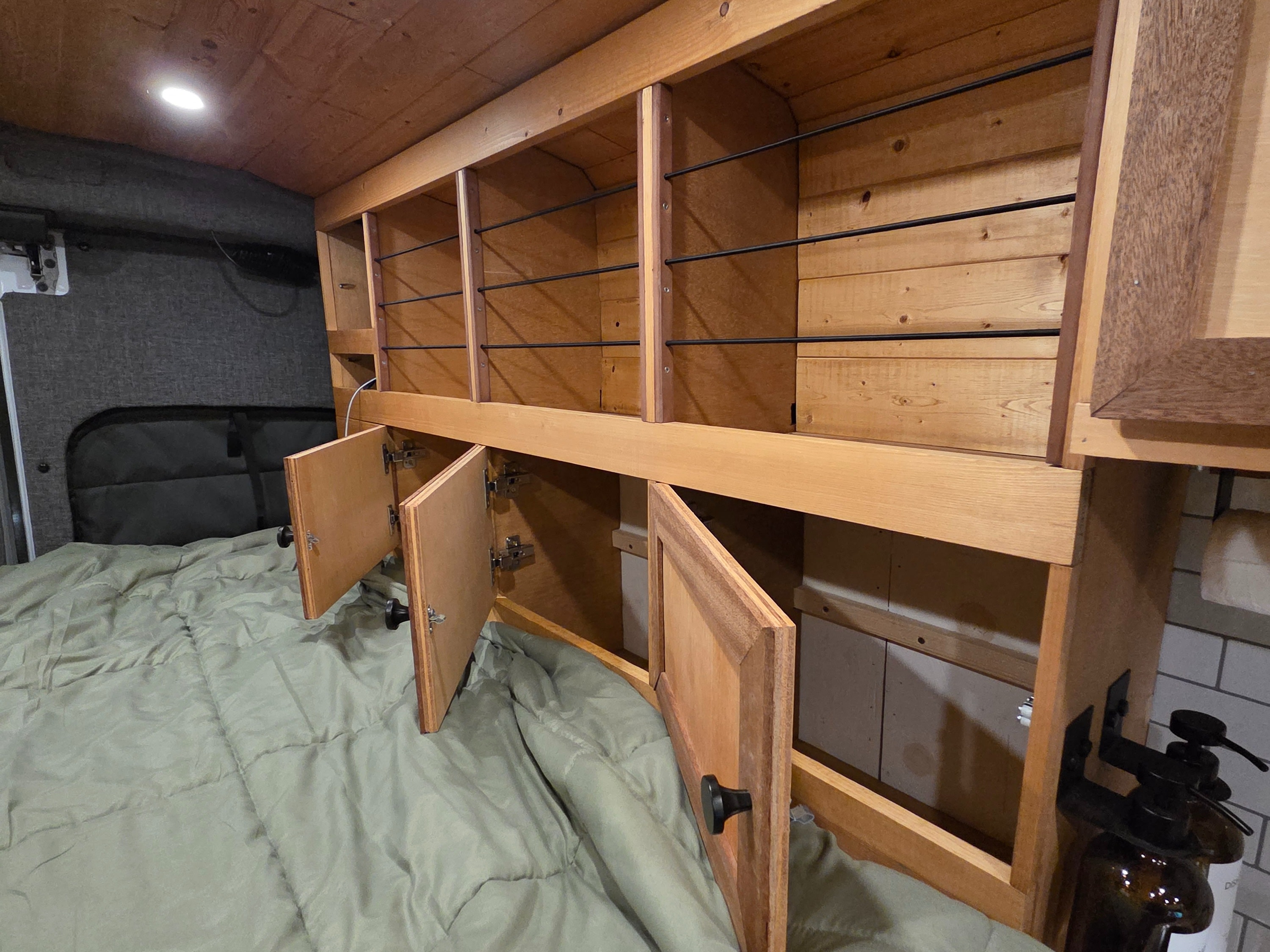 Custom wooden storage and a comfortable bed in an RV rental. Features overhead open shelves and lower cabinets with doors ajar.. Ford Transit 2015